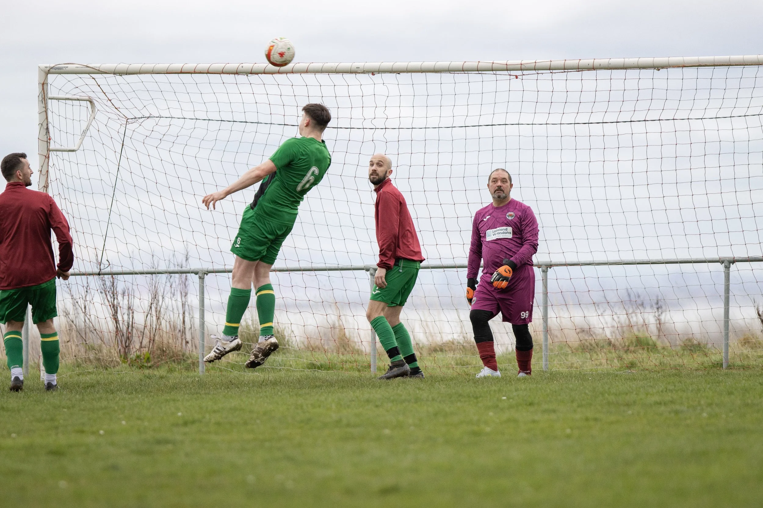 Soccer players on the field with one player heading the ball towards the goal, while others, including the goalkeeper, watch nearby.