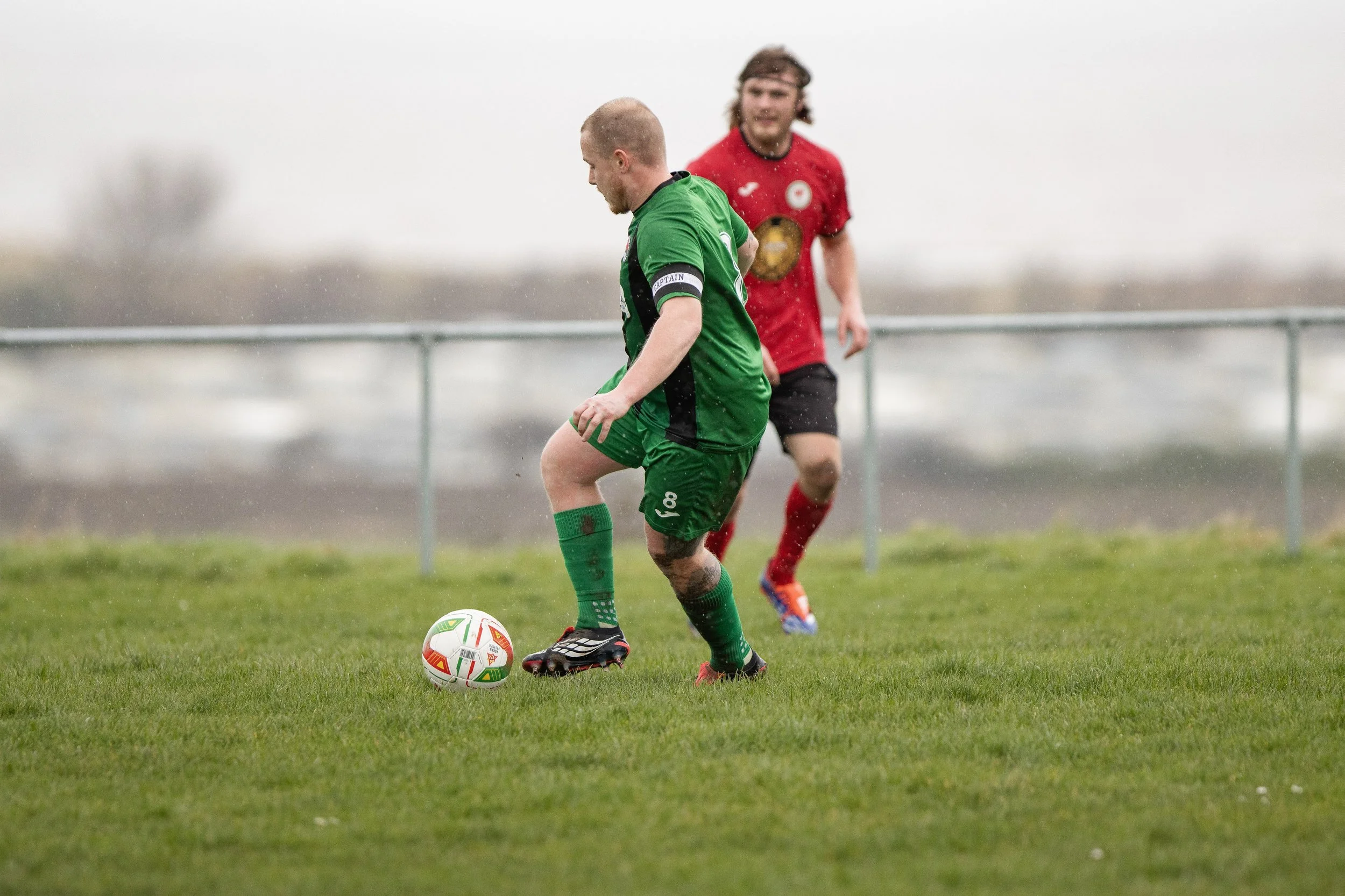 Two soccer players, one in a green uniform and the other in a red uniform, playing on a grassy field during a cloudy day.