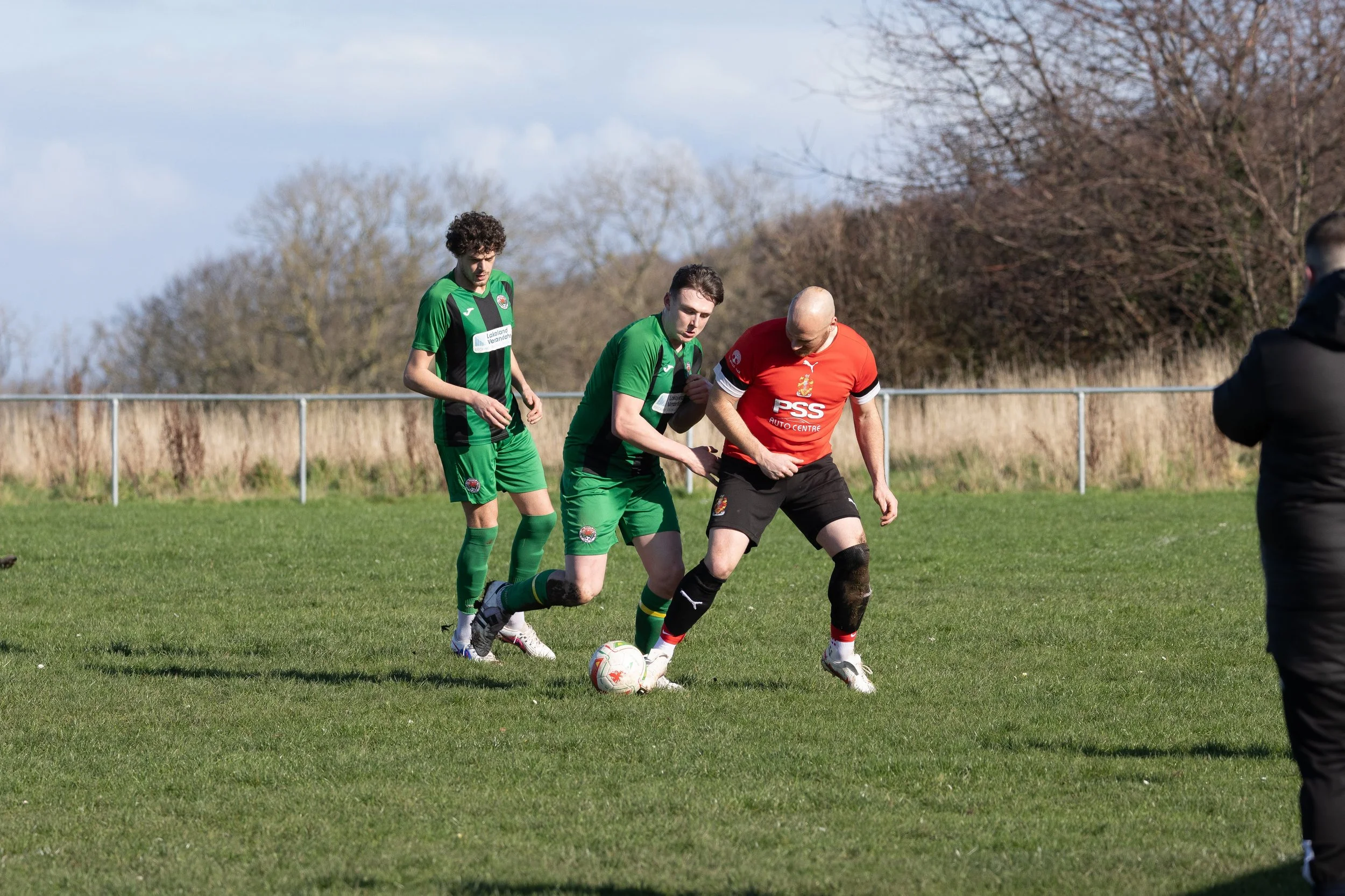 Three soccer players in action on a grassy field, two wearing green jerseys and one in a red jersey, with a person in black on the side and a fence with trees in the background.