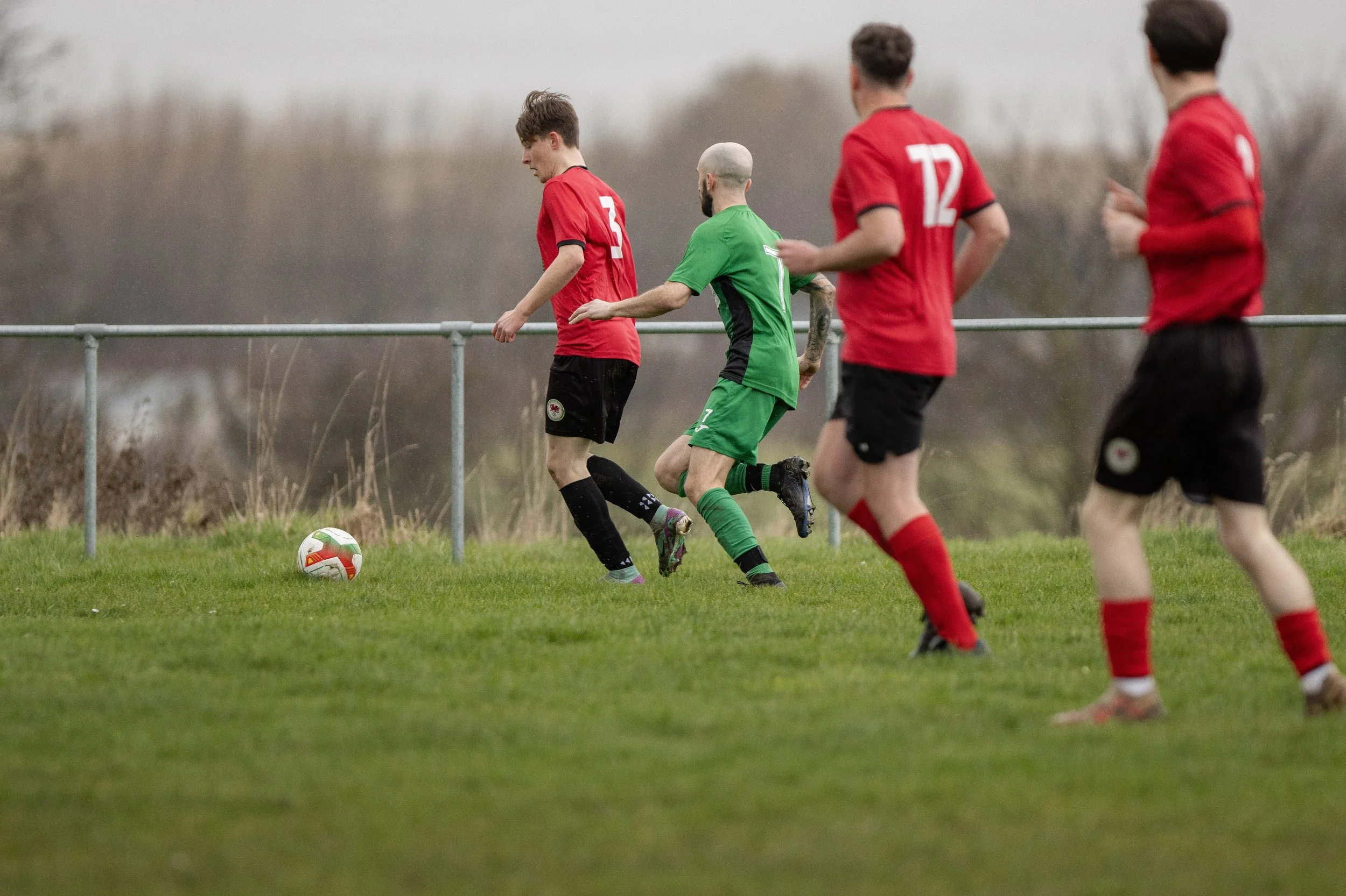 Four soccer players, three in red jerseys and black shorts, and one in a green kit, are playing on a grassy field with a metal railing and trees in the background. The player in green is trying to gain possession of the ball near the sideline.