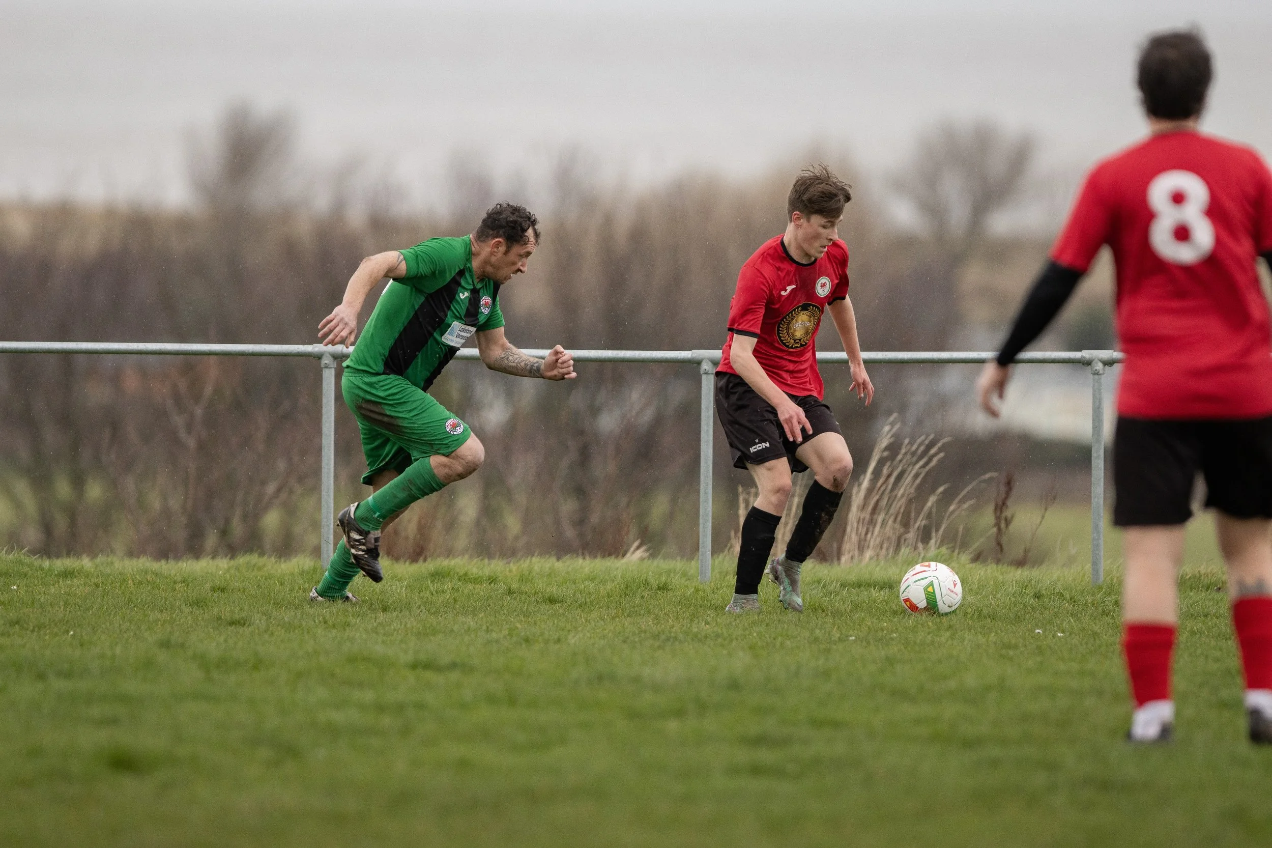 Soccer players in red and green uniforms playing on a grassy field during a match, with two players near the ball and one in the background.