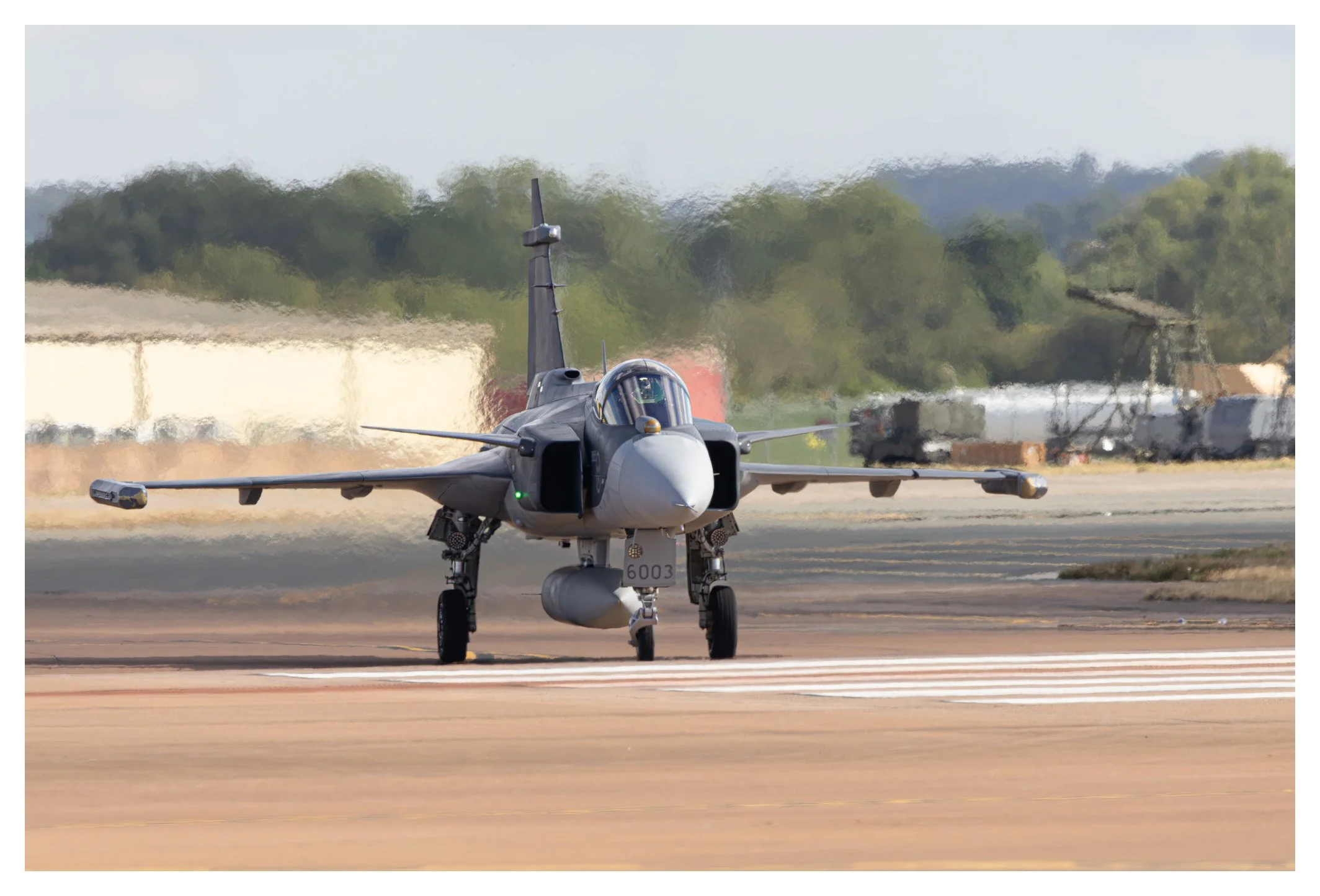 A military fighter jet taxiing on an airport runway with trees and industrial structures in the background.