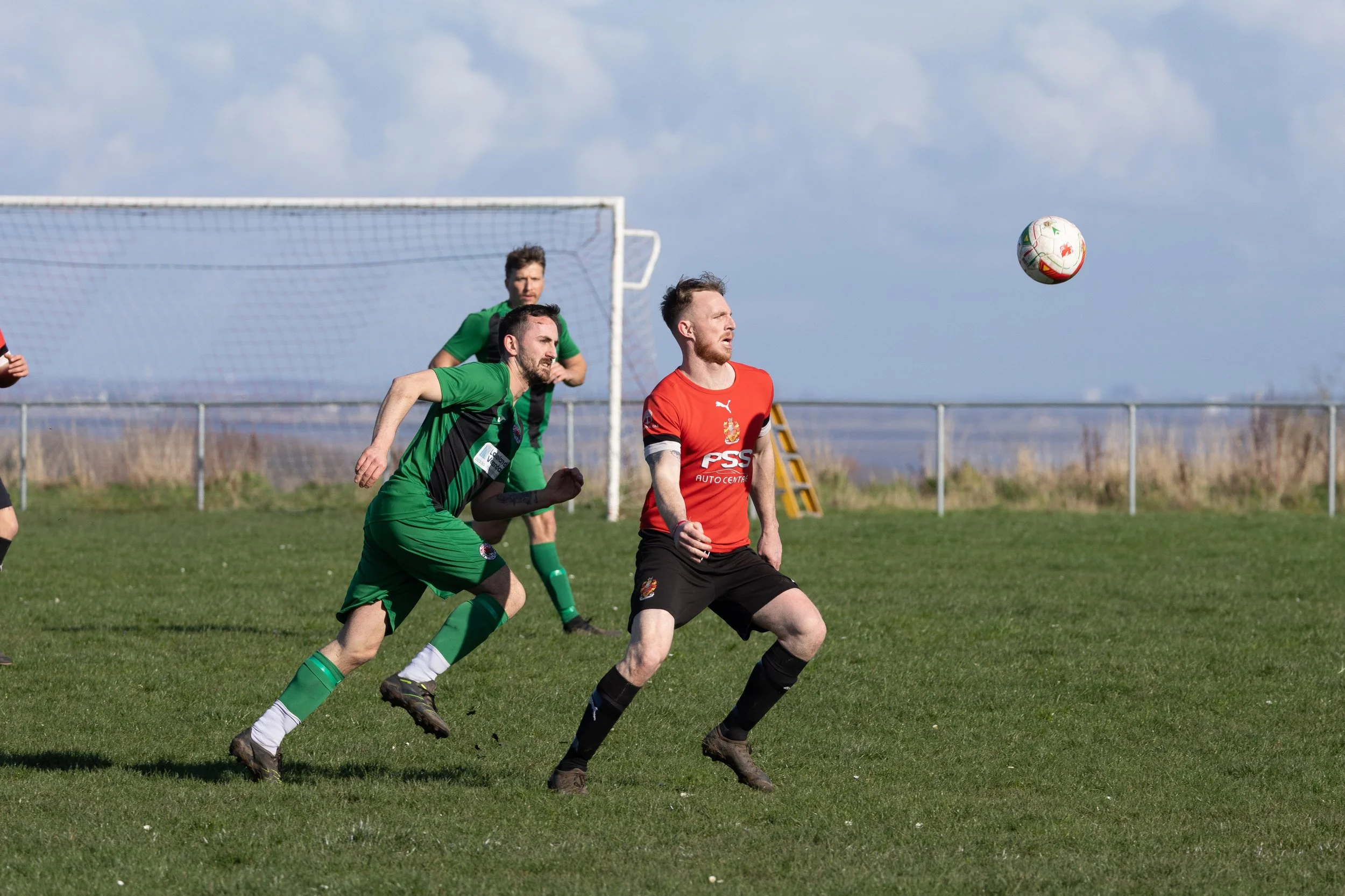 Soccer players in action on the field, with one in a red jersey and two in green jerseys, chasing the ball near the goalpost under a blue sky with clouds.