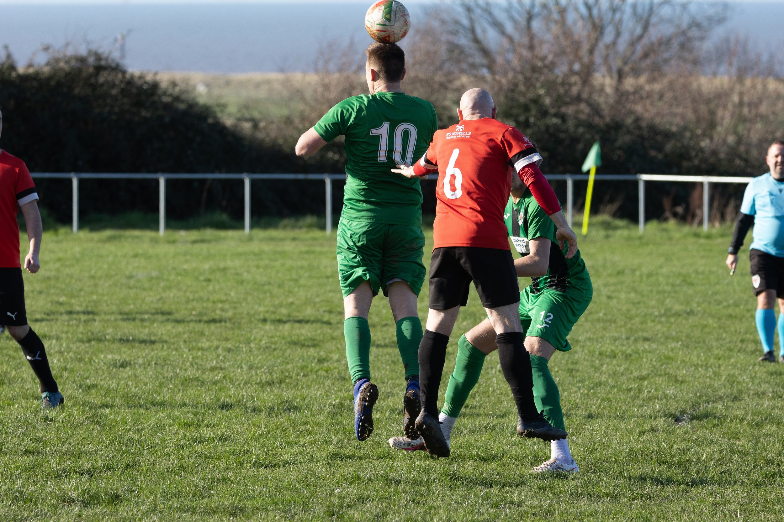 Soccer players in green and red uniforms compete for the ball in the air during a match on a grassy field, with a blue sky and trees in the background.