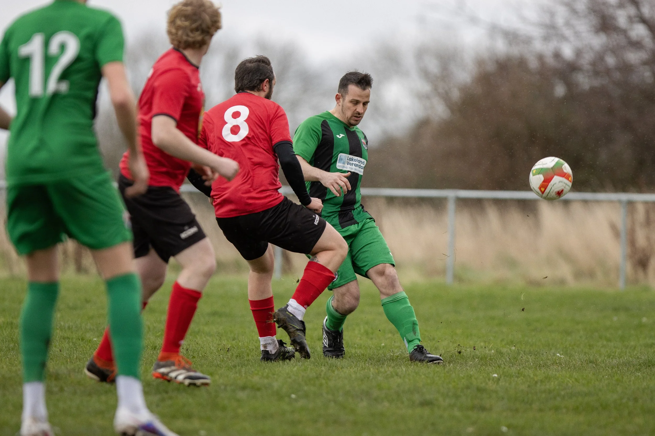Soccer players competing for the ball on the field during a match, with one player in a green and black uniform and others in red and black uniforms, on a grassy field with a metal fence and trees in the background.