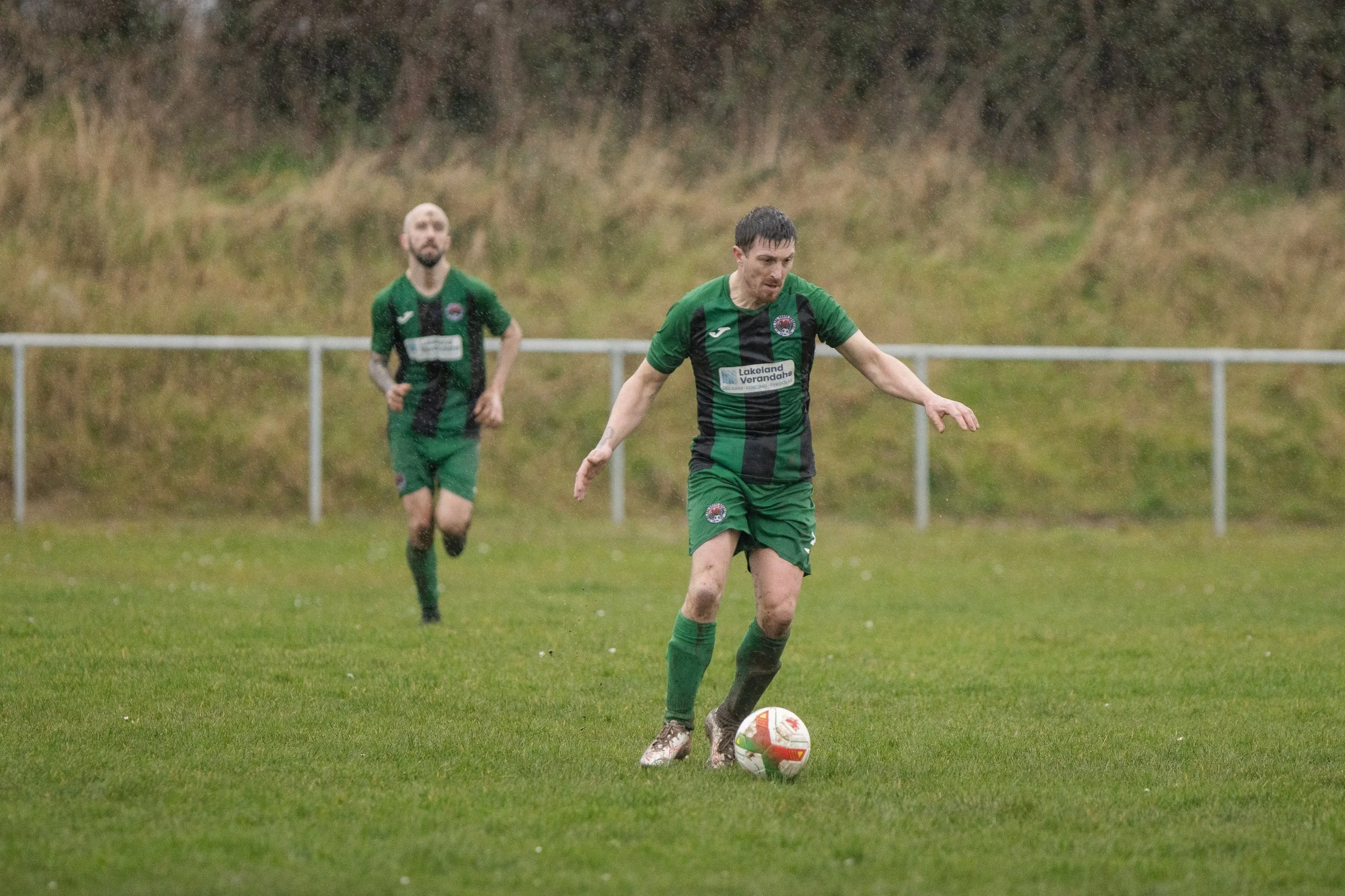 Two male soccer players in green and black uniforms playing on a grass field in rainy weather, with one player preparing to kick a soccer ball and the other running behind him.