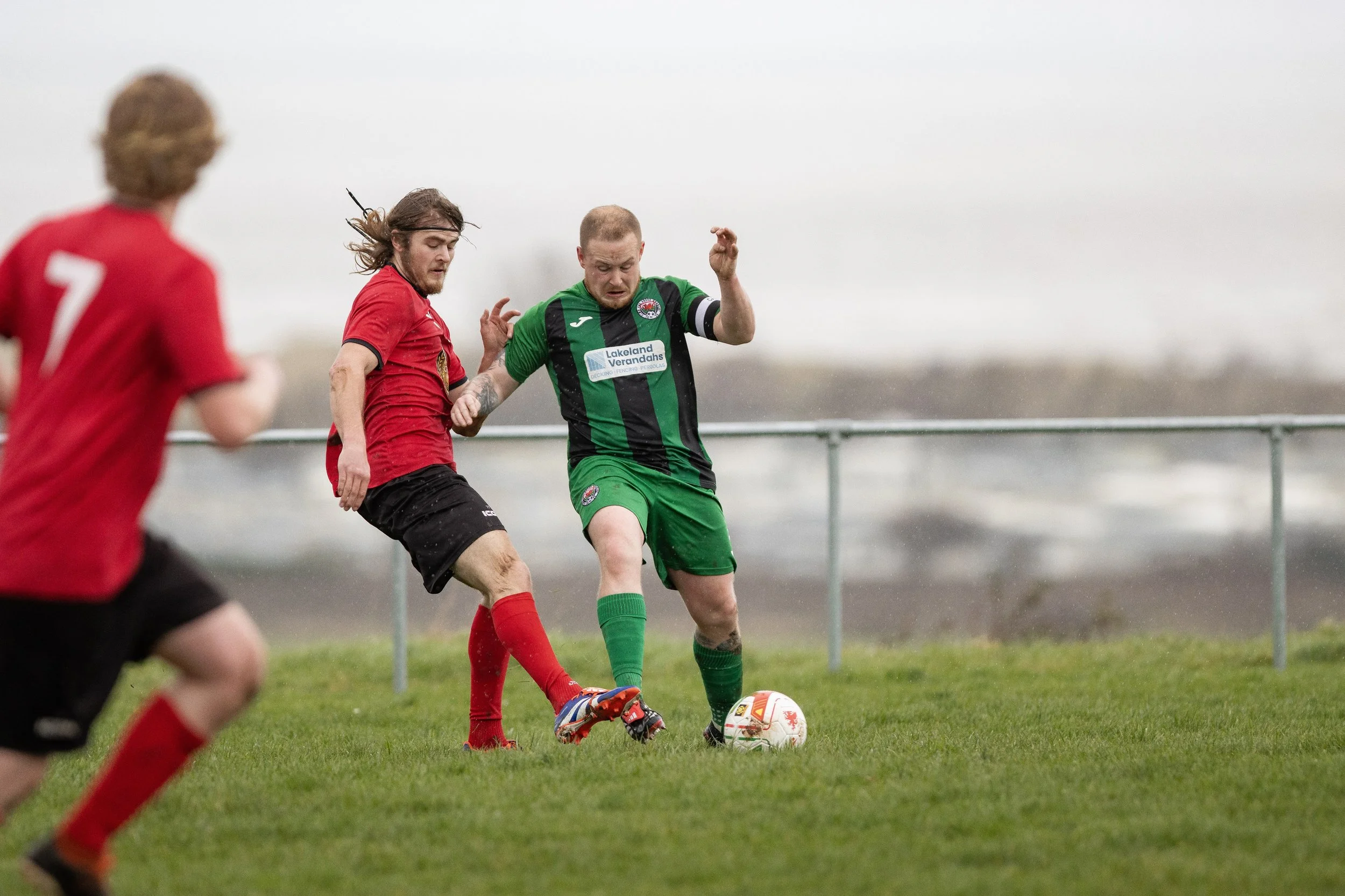 Two soccer players, one in a red jersey and black shorts, the other in a green and black jersey and green shorts, compete for the ball on a grassy field during a game, with a third player in red partially visible in the foreground.