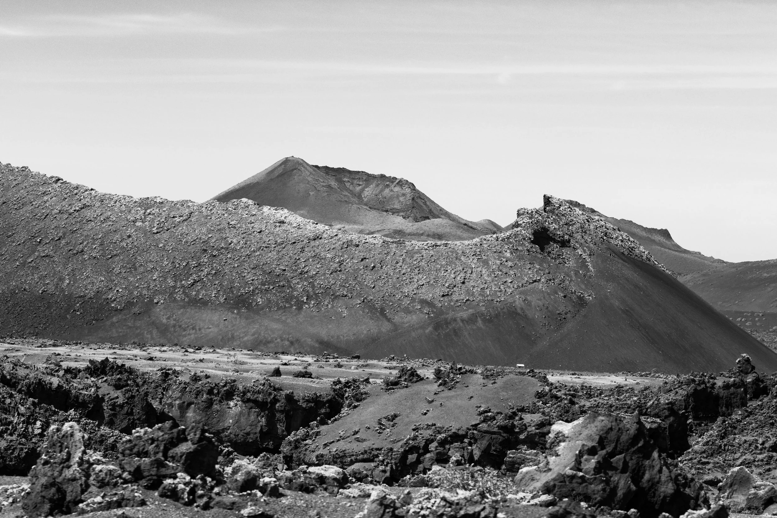 Black and white photo of a volcanic mountain range with rugged terrain, rocky slopes, and visible volcanic craters under a cloudy sky.