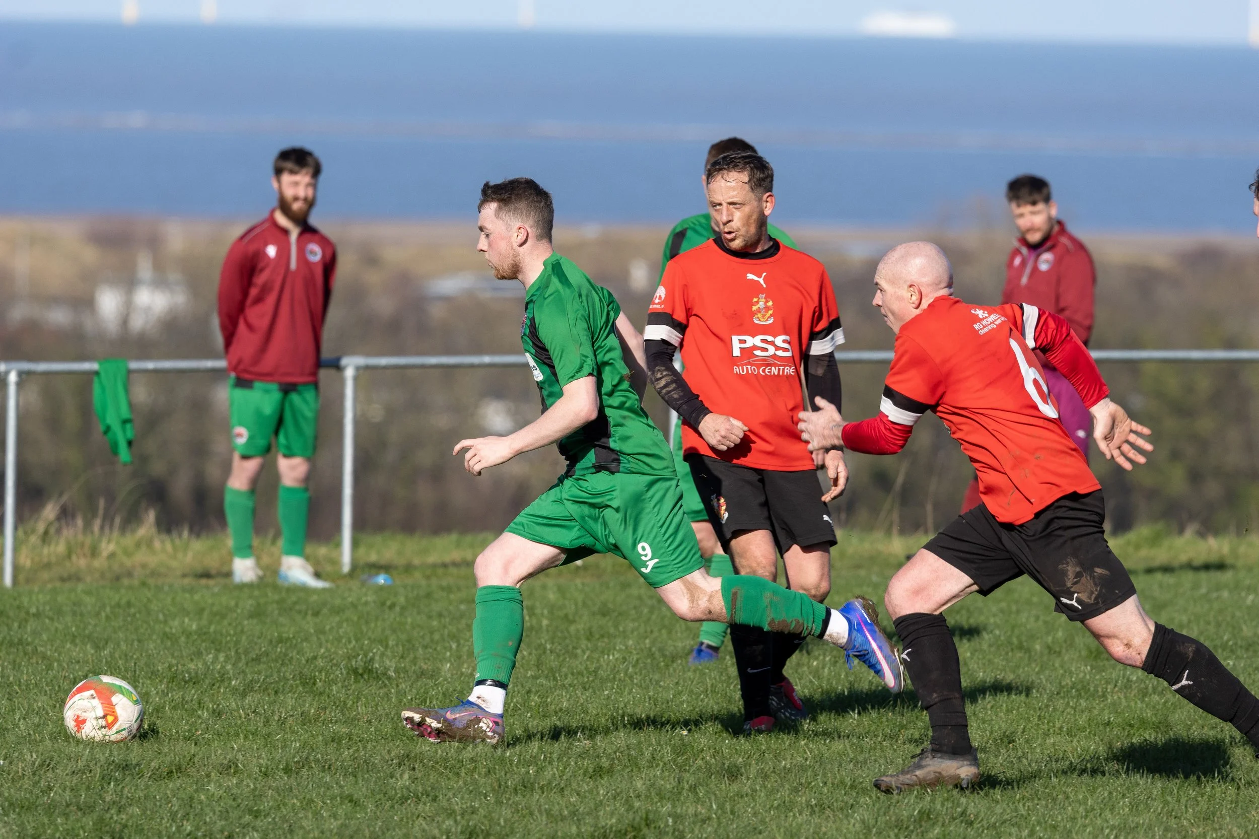 Soccer players in red and green jerseys competing for the ball on a grassy field with a few people watching in the background.