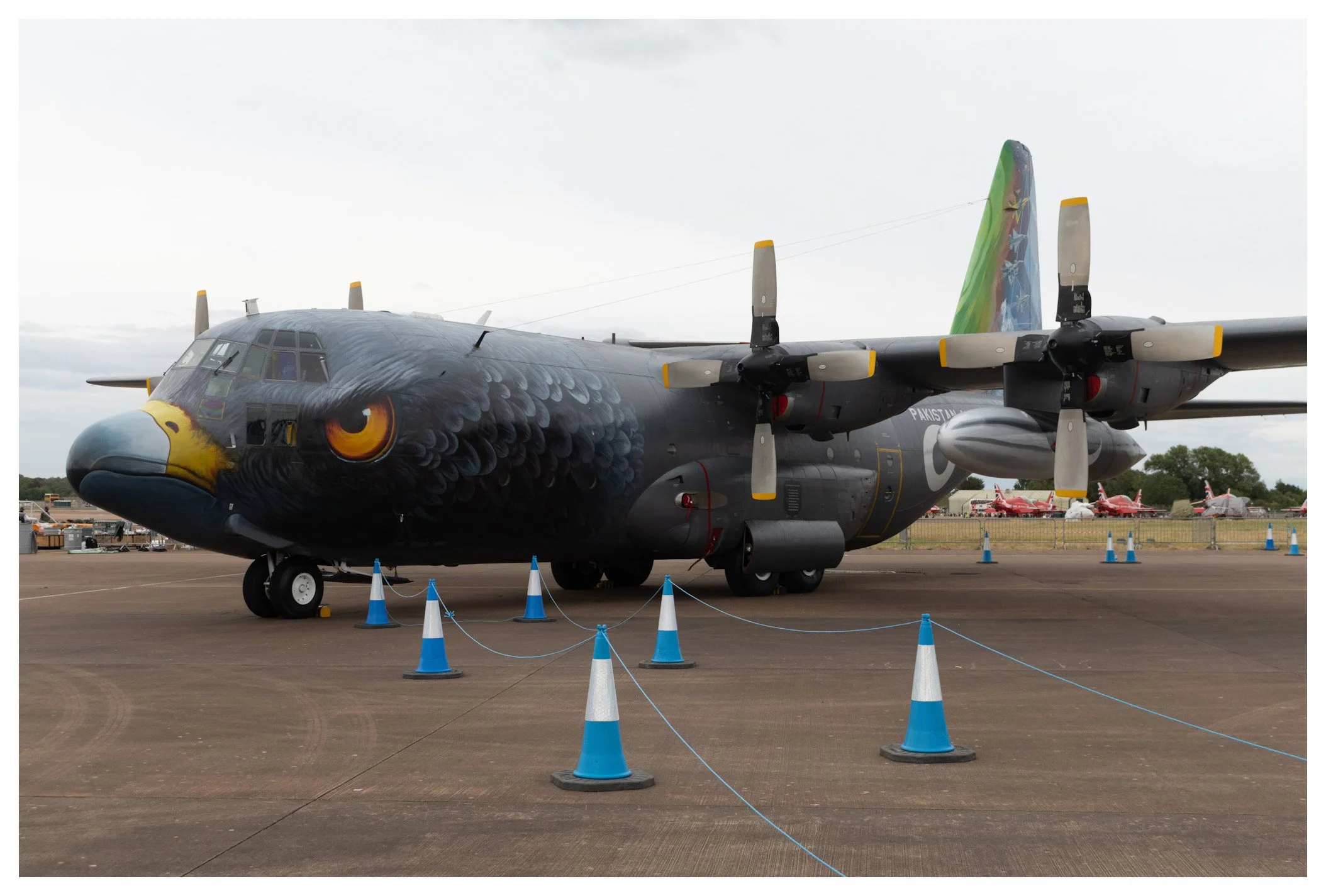 A military transport airplane with an eagle face painted on the nose and an eagle design on the tail, parked on a tarmac with blue cones around it.