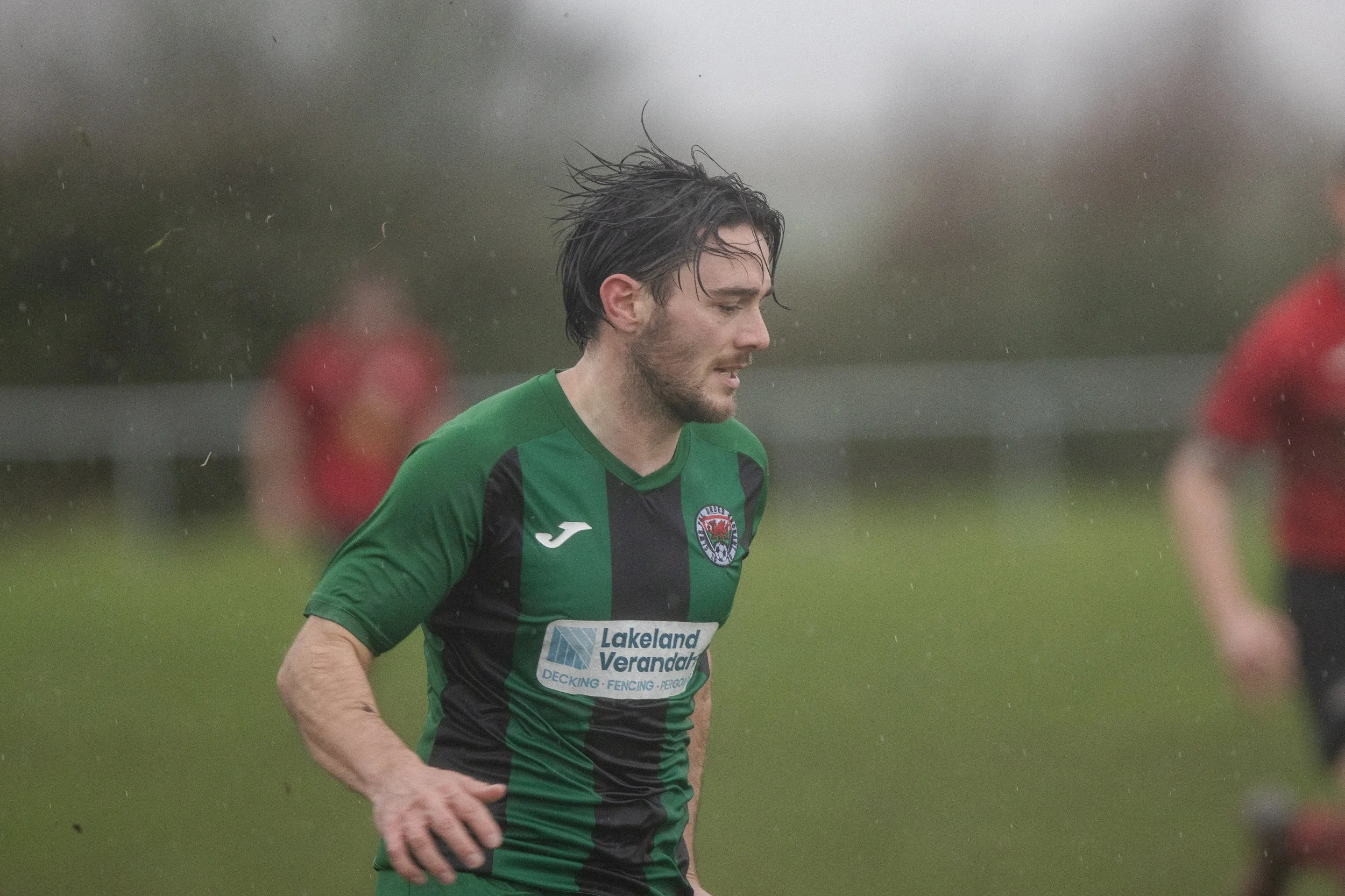 A soccer player in green and black uniform playing in rain, with other players blurred in the background.