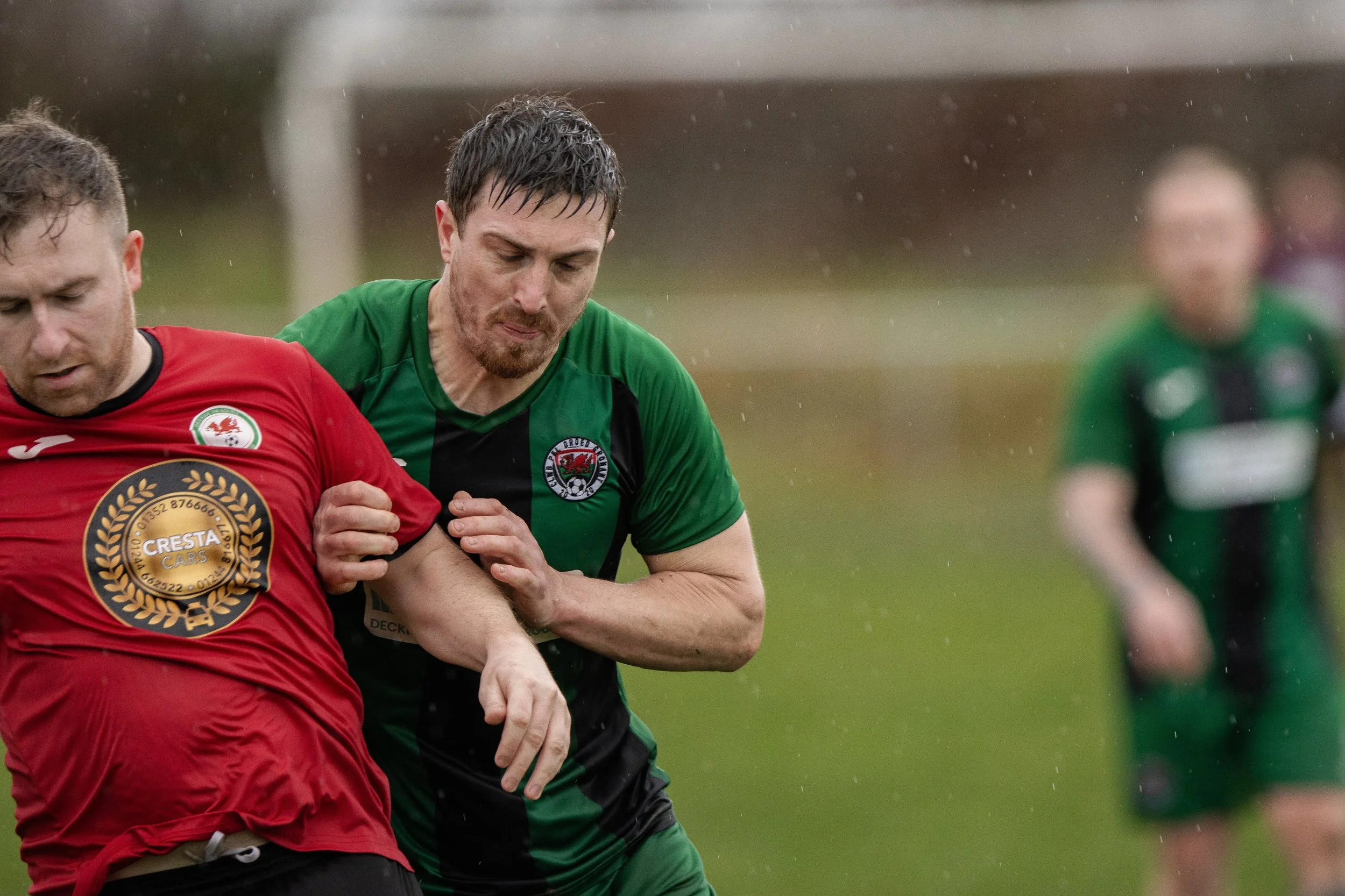 Two rugby players, one in a red jersey and one in a green and black jersey, contest for the ball in a rainy match. A third player in a green and black jersey is visible in the background.