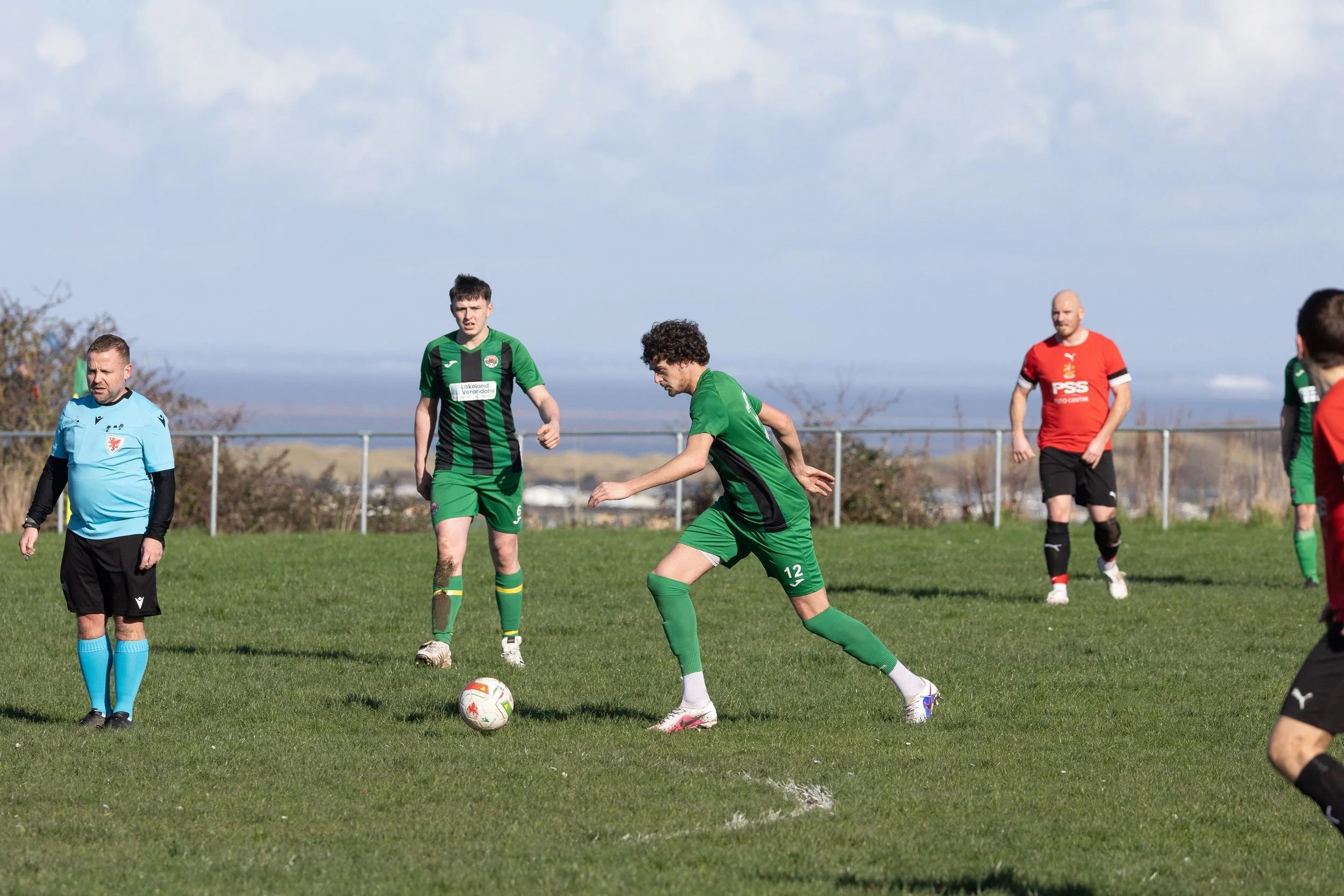 Soccer players on the field during a match, with a player in a green jersey in the center preparing to kick the ball, surrounded by other players and a referee in a light blue shirt, under a partly cloudy sky.