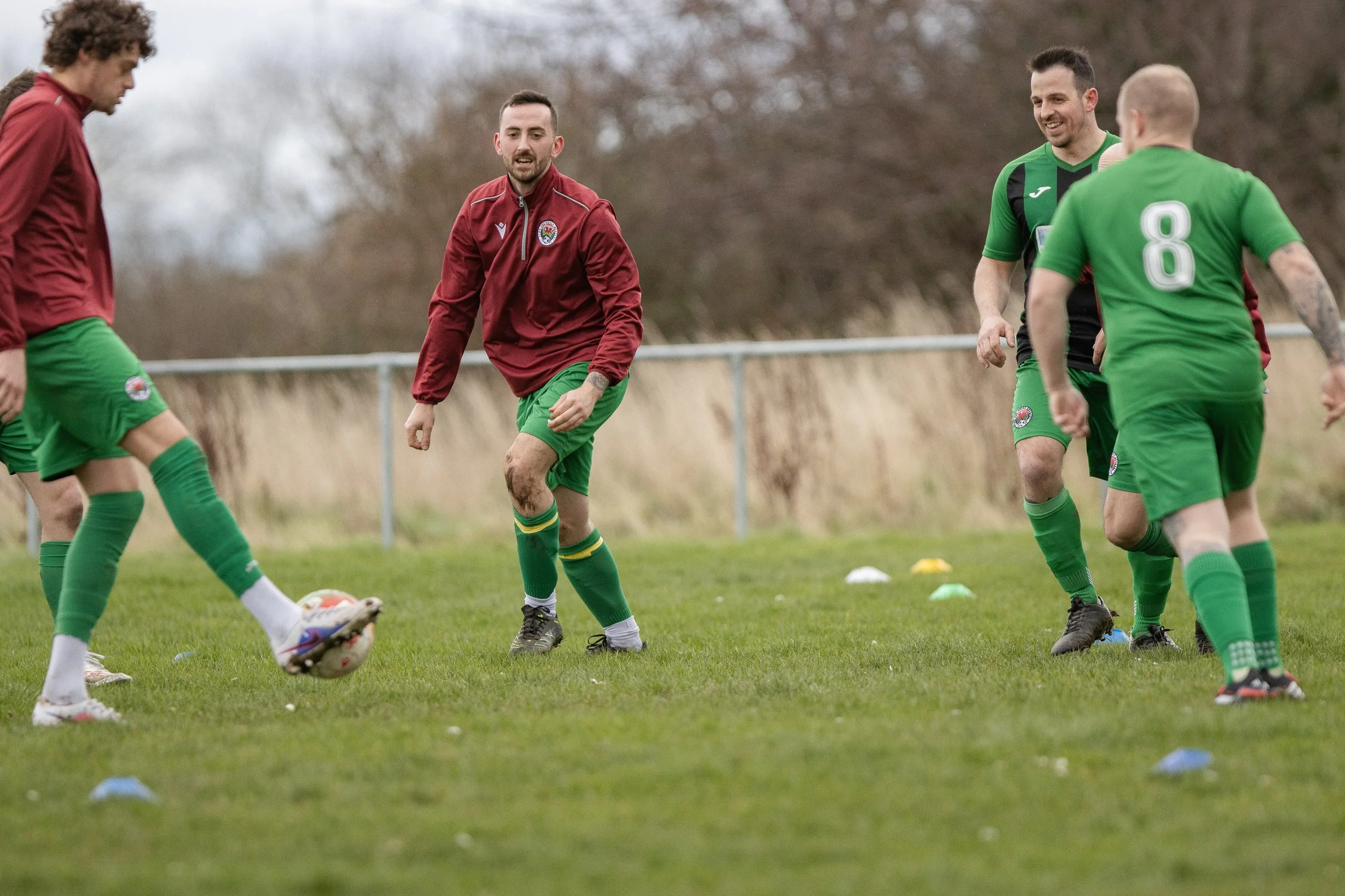 Soccer players practicing on a grassy field with cones, wearing green and maroon uniforms, with a fence and trees in the background.