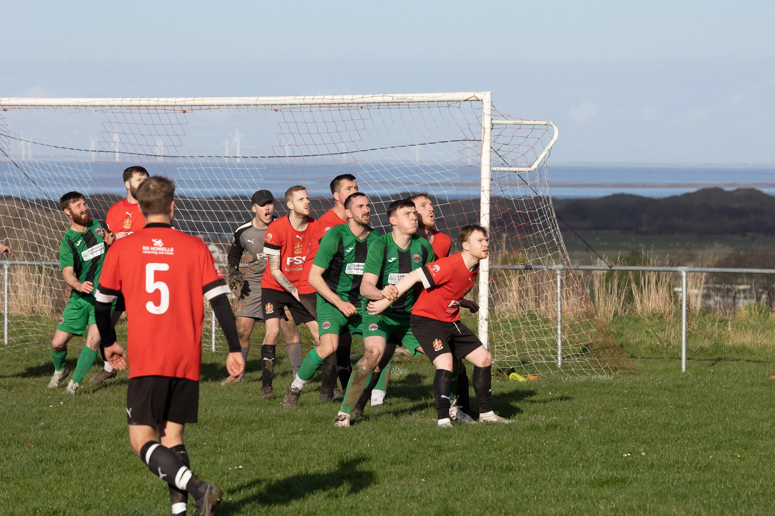 Soccer players in red and green jerseys defending their goal during a match on a grassy field.