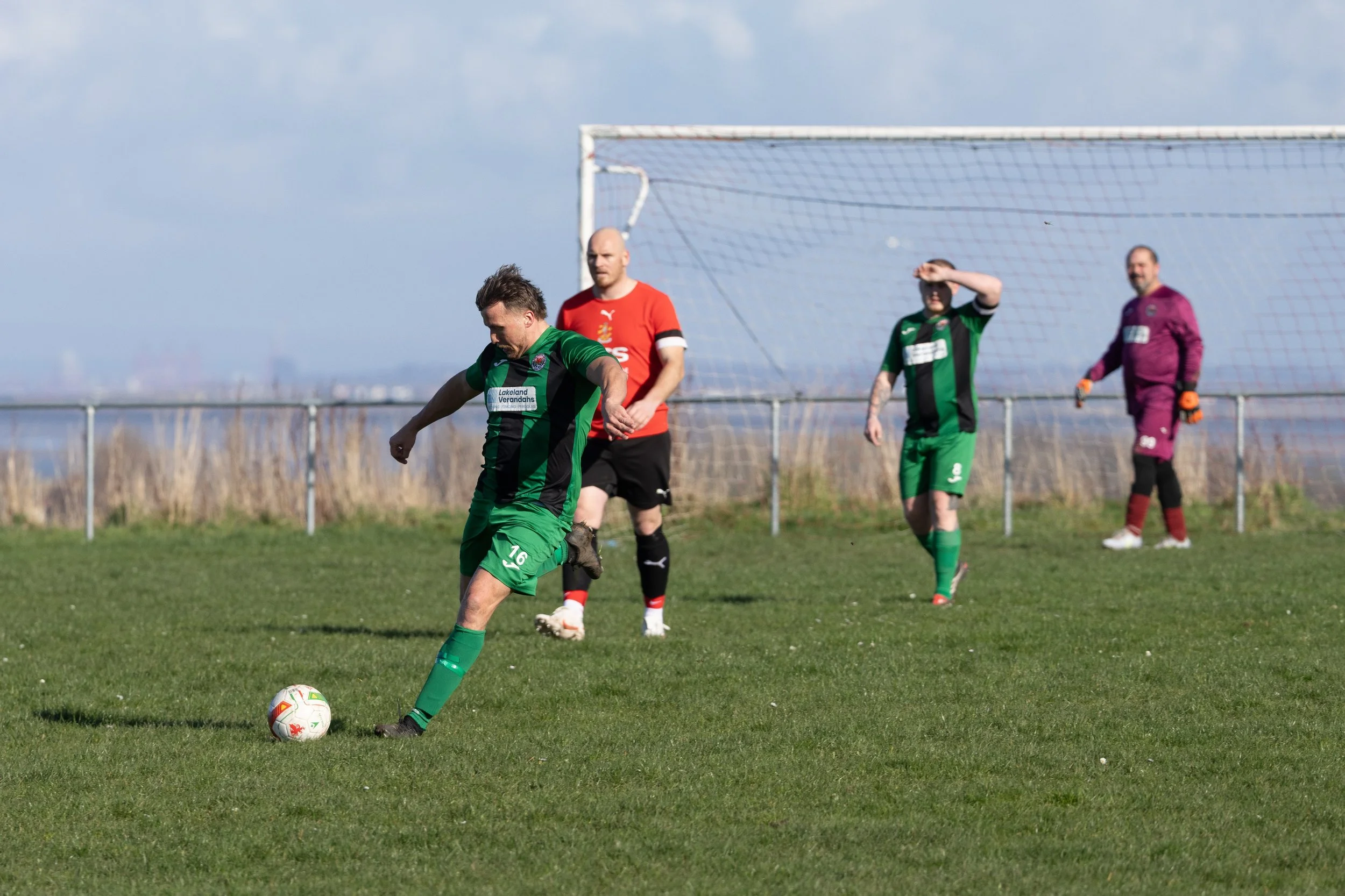 Soccer players on a grassy field with a goalpost in the background. One player in green is preparing to kick the ball, while others, including a goalkeeper in purple, stand nearby.