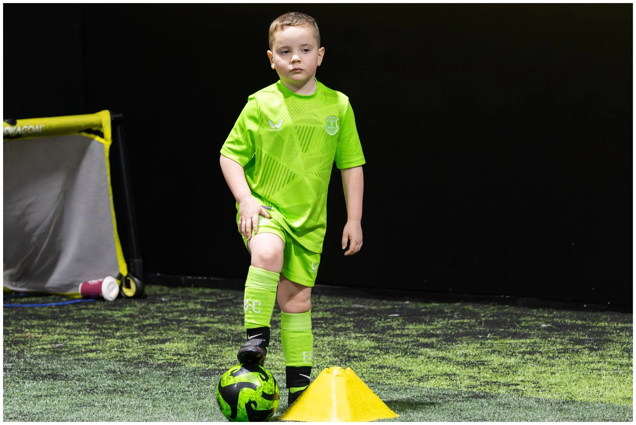 Young boy in bright green soccer uniform standing on indoor field, with one foot on a green and black soccer ball, next to a yellow training cone, in front of a dark background.