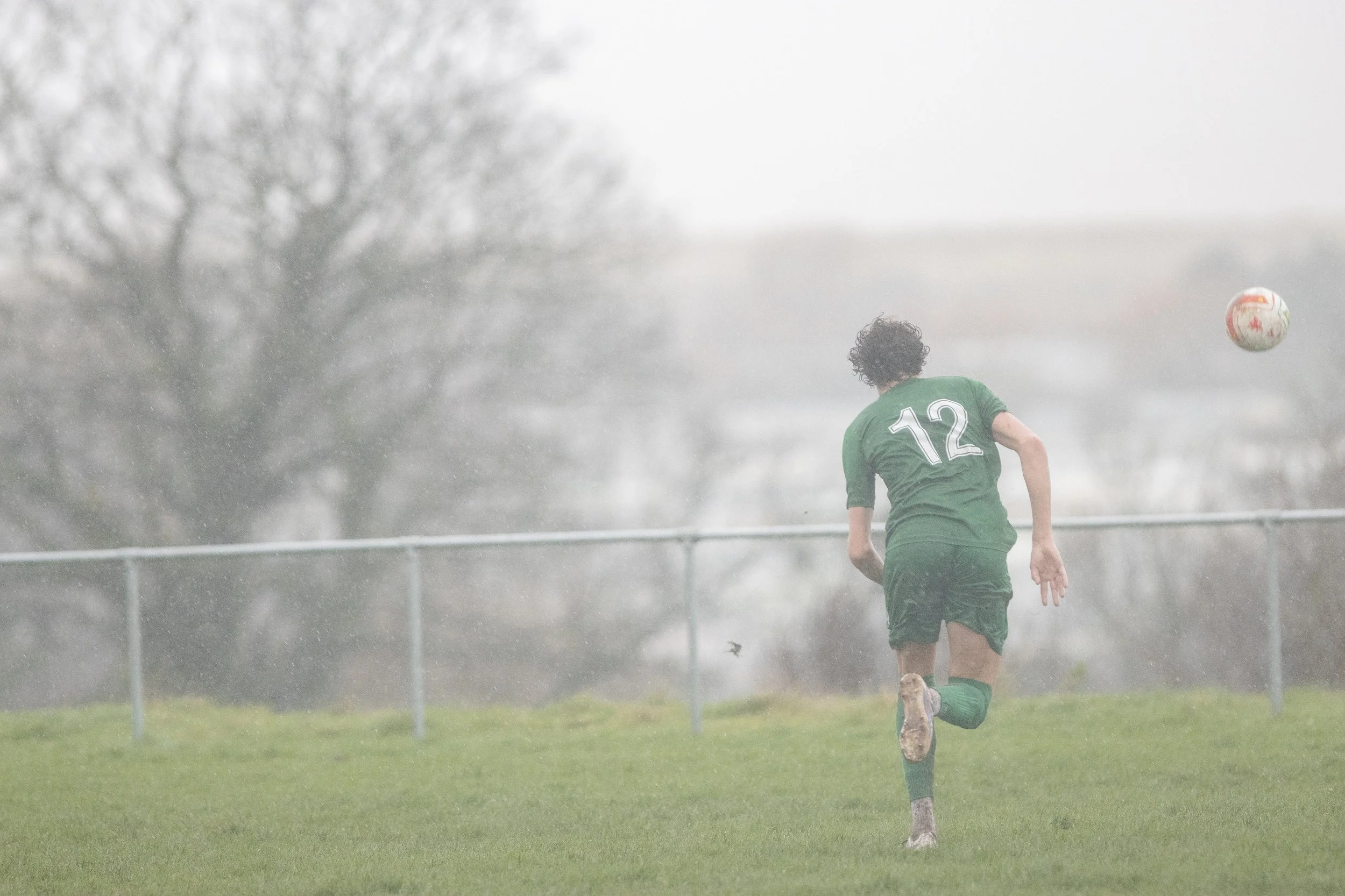A soccer player in a green uniform with the number 12 on the back runs on a wet, grassy field during rainy weather, with a soccer ball in the air nearby.