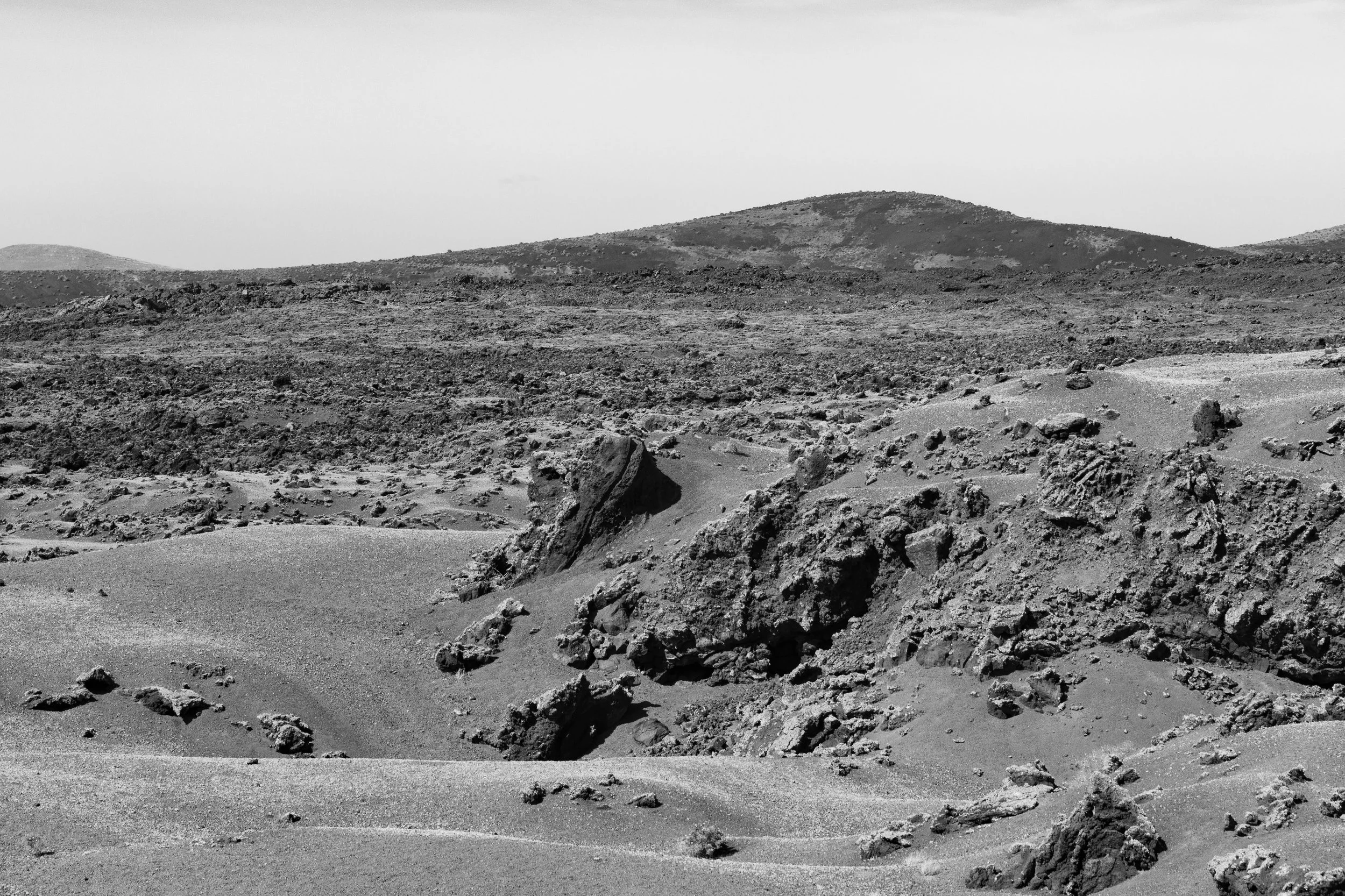 Black and white photograph of a barren, rocky desert landscape with sand dunes and distant hills under clear sky.