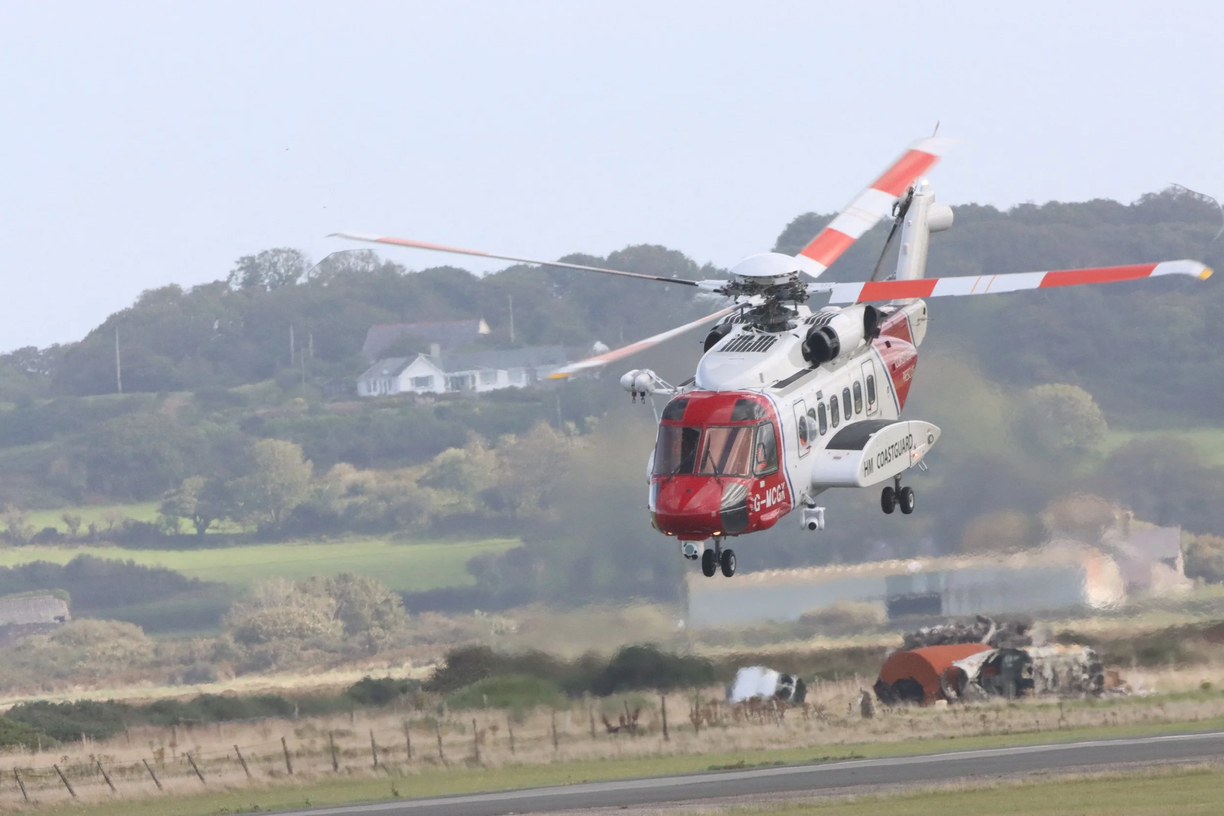A helicopter flying low over a grassy field with wreckage and a burnt vehicle on the ground, and houses on a hill in the background.