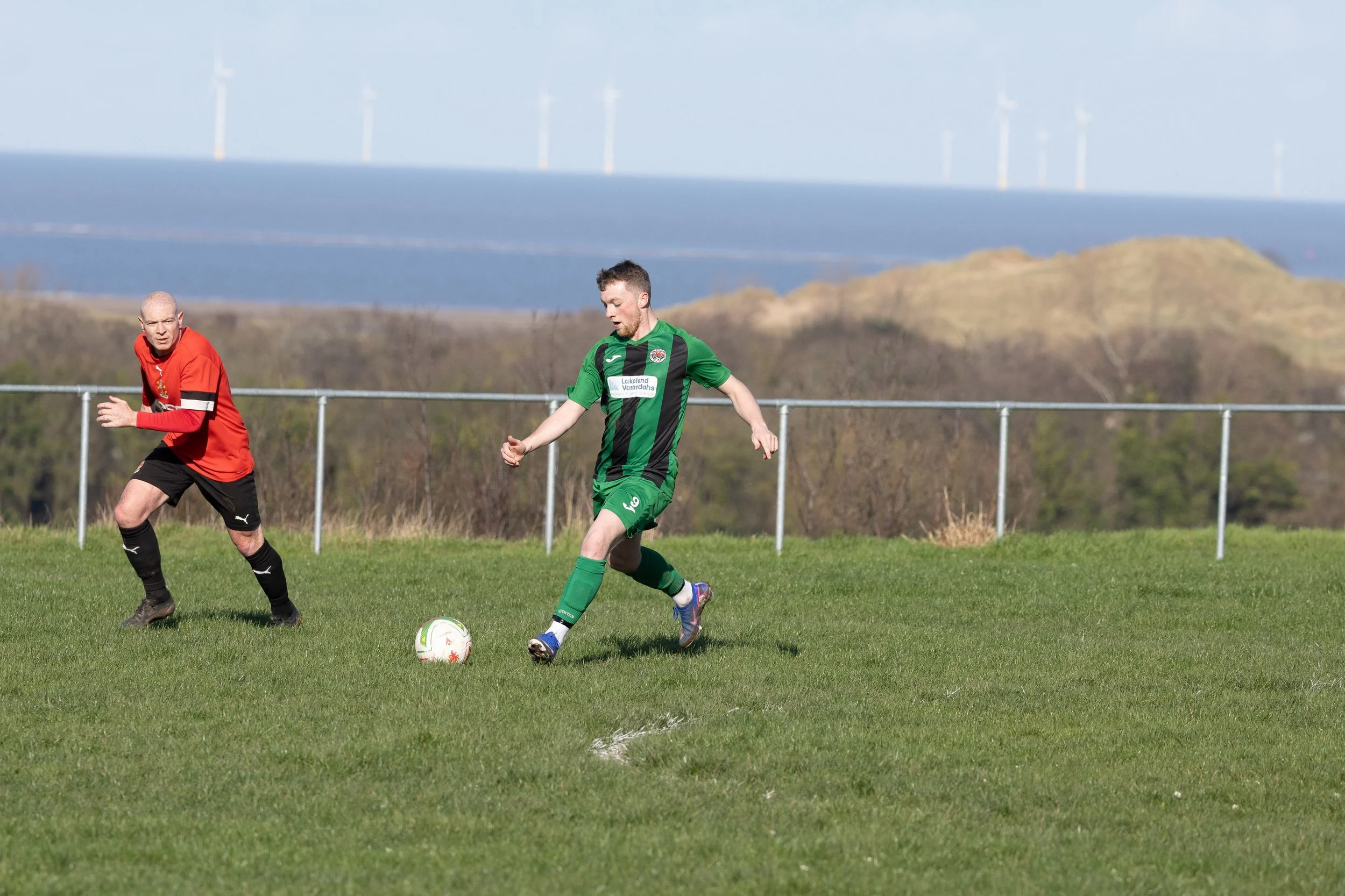Two soccer players on a grassy field, one in a green uniform and the other in a red and black uniform, with a coastal landscape and wind turbines in the background.