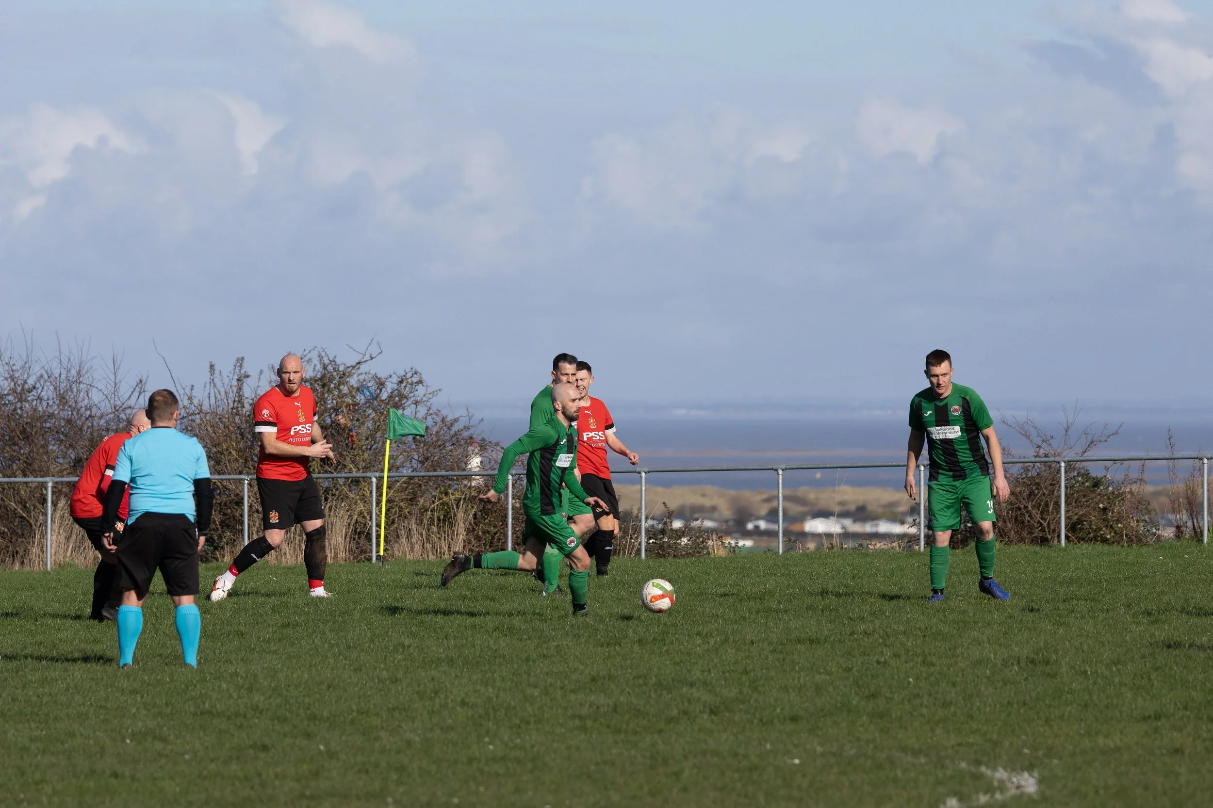 Soccer players on the field during a game, with one player in green preparing to kick the ball, others in red and a referee in blue nearby, on a grassy field with a fence and a cloudy sky in the background.