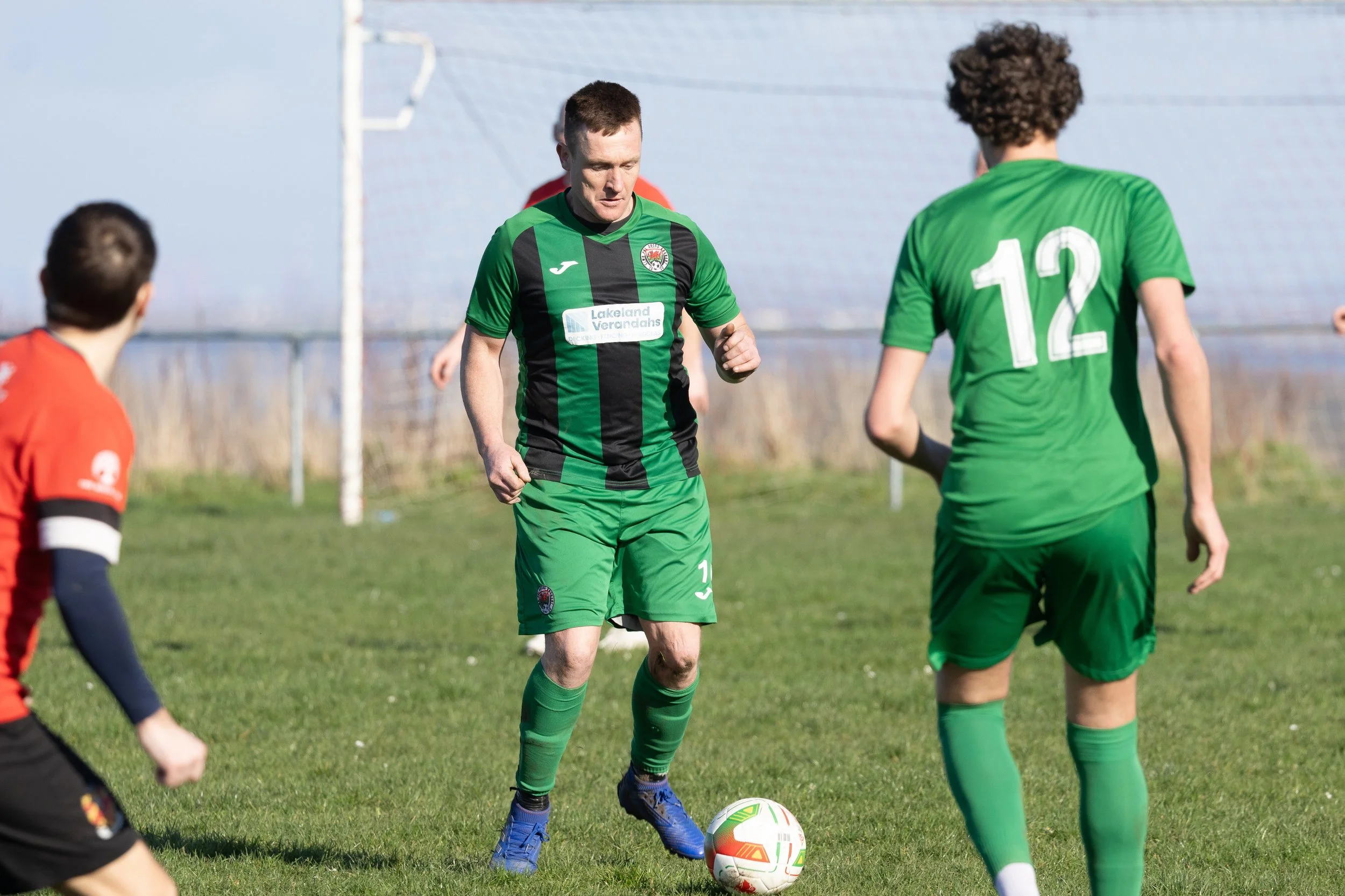 A soccer game in progress with a player in a black and green uniform about to kick the ball, surrounded by players in green and red uniforms on a grassy field.