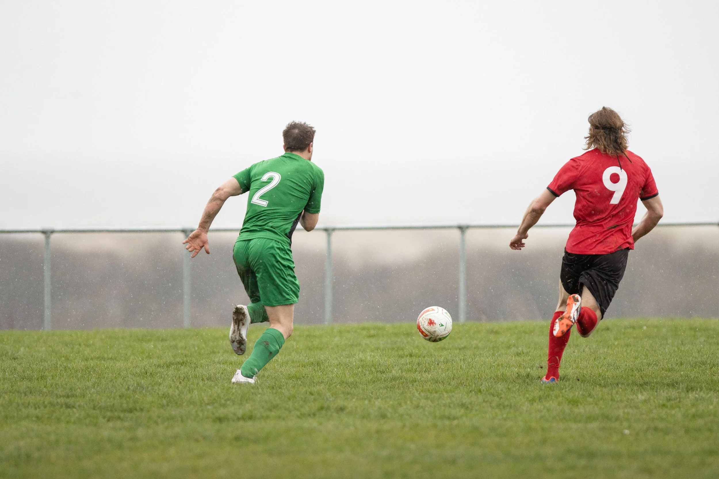 Two soccer players running towards a soccer ball on a grassy field, one in a green uniform with the number 2, and the other in a red uniform with the number 9, during a game.