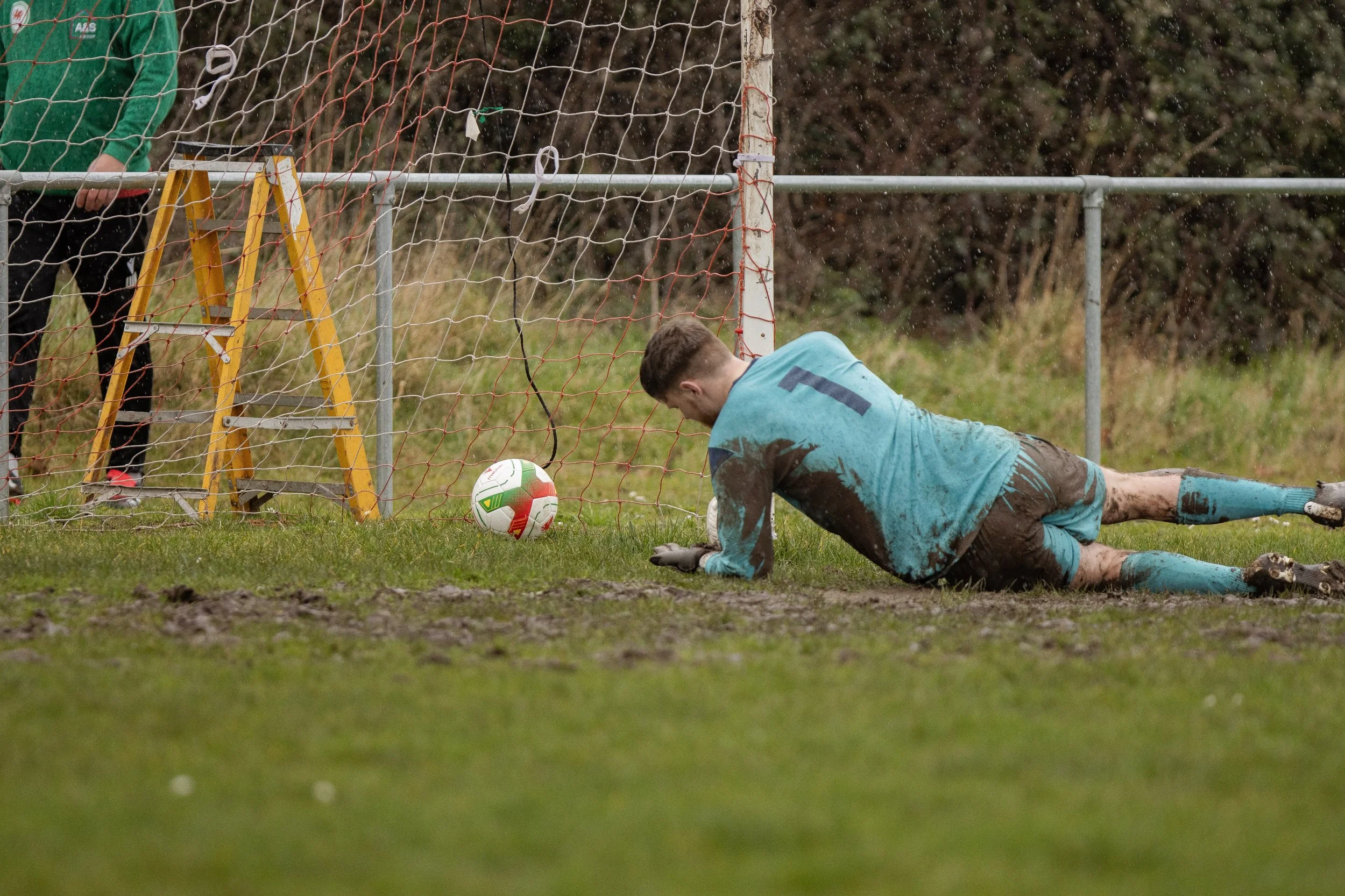 A soccer goalie wearing a blue uniform with the number 1 on the back lying on the muddy field, after failing to stop a soccer ball near the goalpost. The goal net and a person on a yellow ladder are visible in the background.