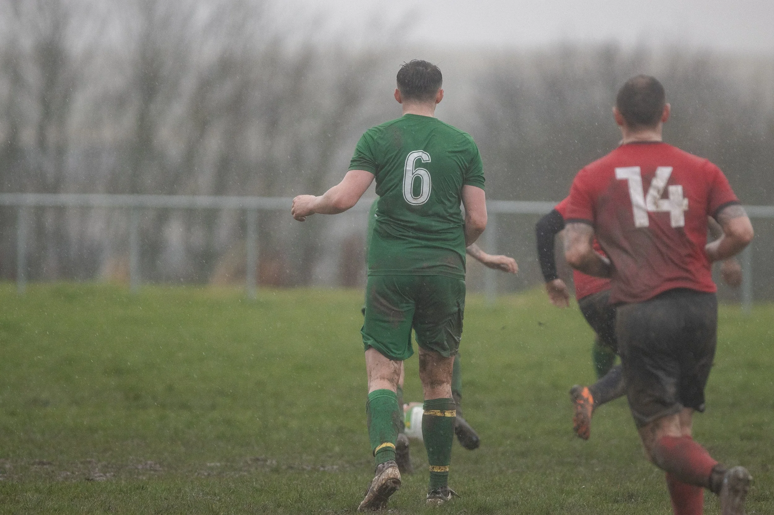 Soccer players in rainy weather on a muddy field, with one player wearing a green jersey with the number 6 and another in a red jersey with number 14.