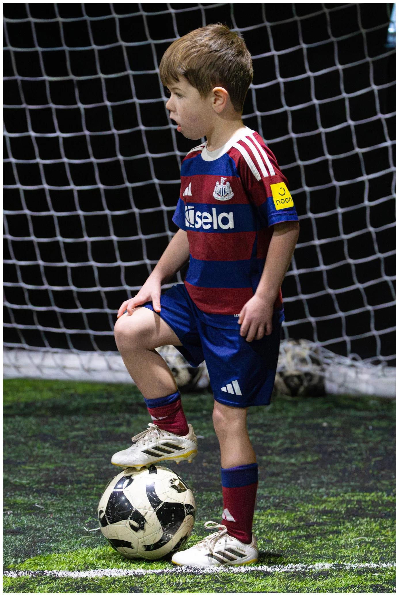 A young boy in a soccer uniform standing on a soccer field at night, with one foot on a soccer ball and a goal net in the background.