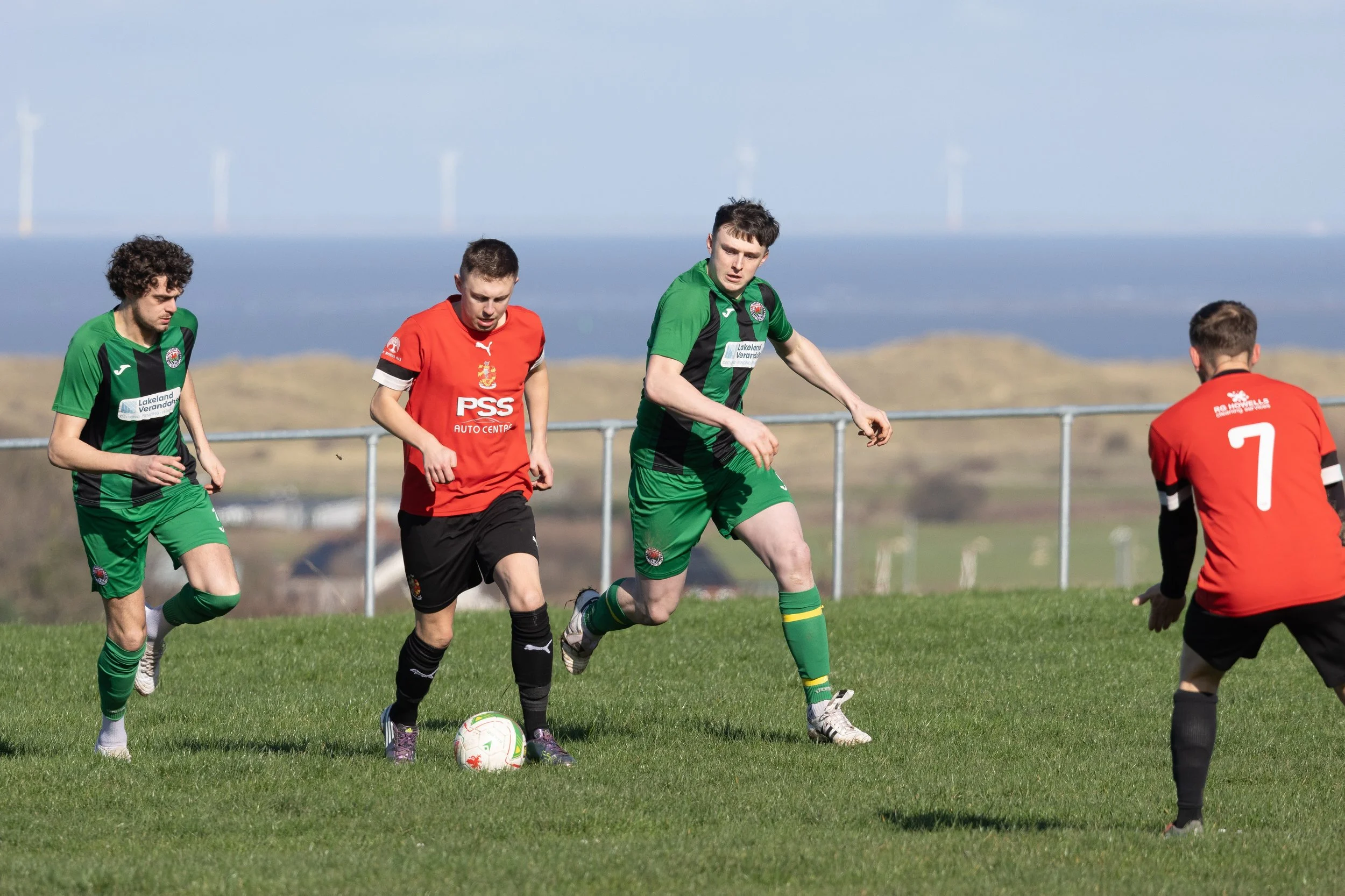 Soccer match with four players on the field, two in green uniforms and two in red uniforms, near a soccer ball on the grass, with a scenic background of open land and wind turbines.