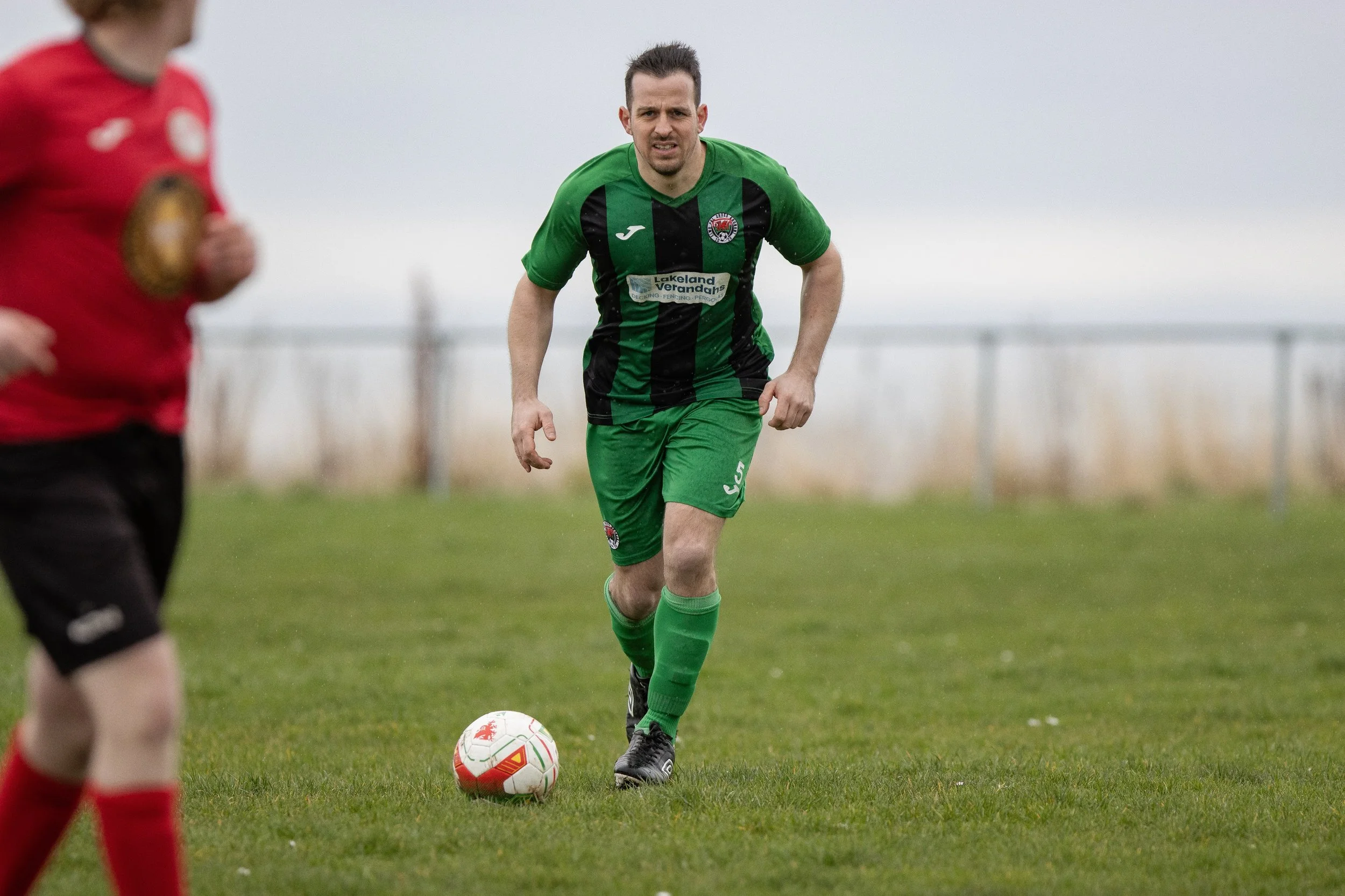 A man in a green and black soccer uniform running on a grassy field with a ball at his feet, with another player partially visible in a red and black uniform in the foreground.