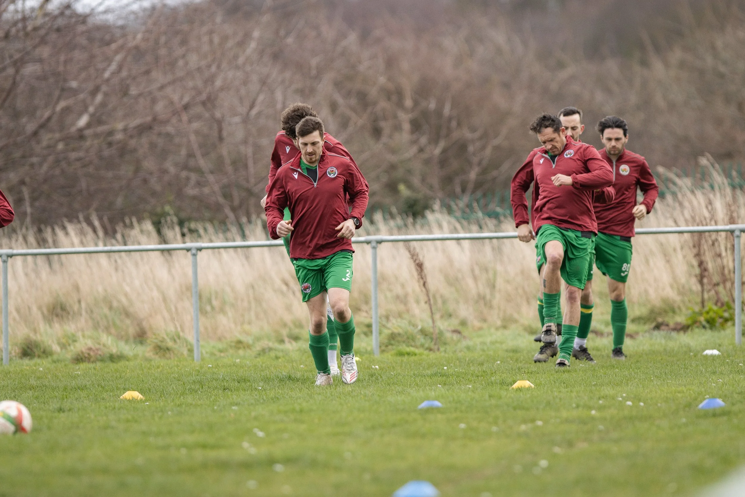 A group of soccer players wearing maroon jackets and green shorts training on a grassy field with cones, during an overcast day.