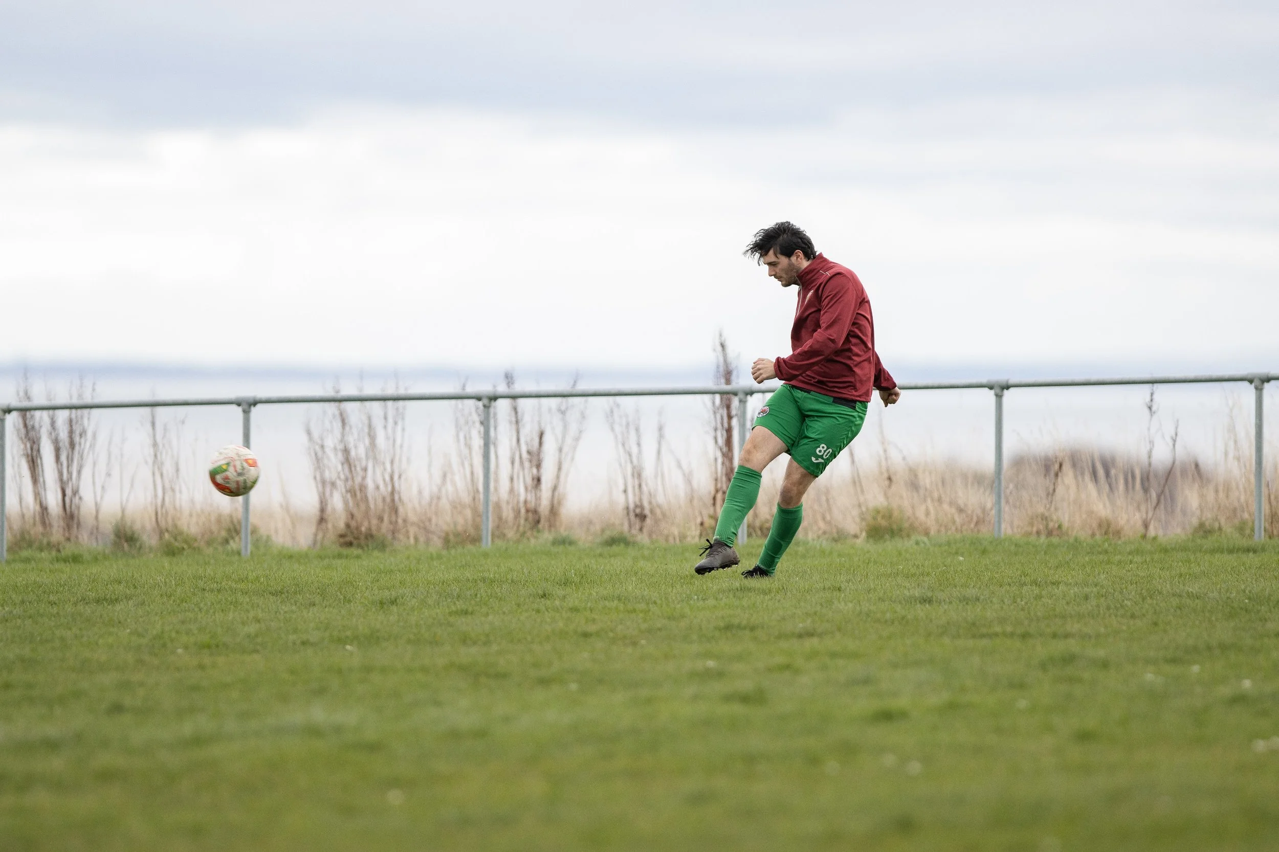A man in a red jacket and green soccer uniform kicking a ball on a grassy field.