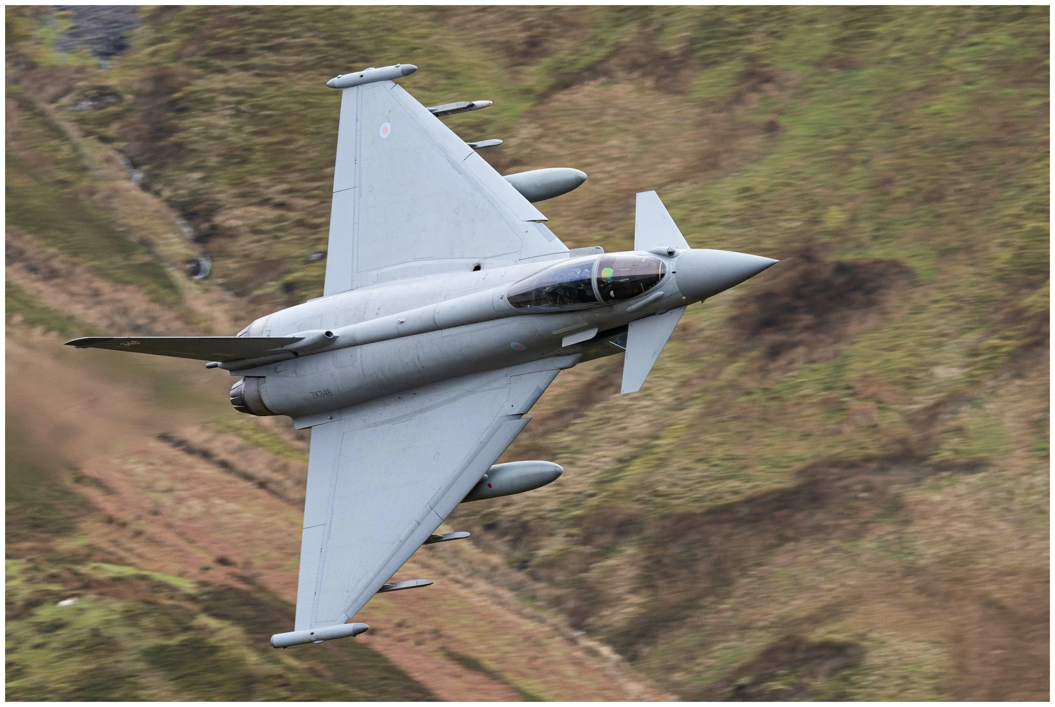 A gray fighter jet aircraft flying at an angle with a mountainous landscape in the background.