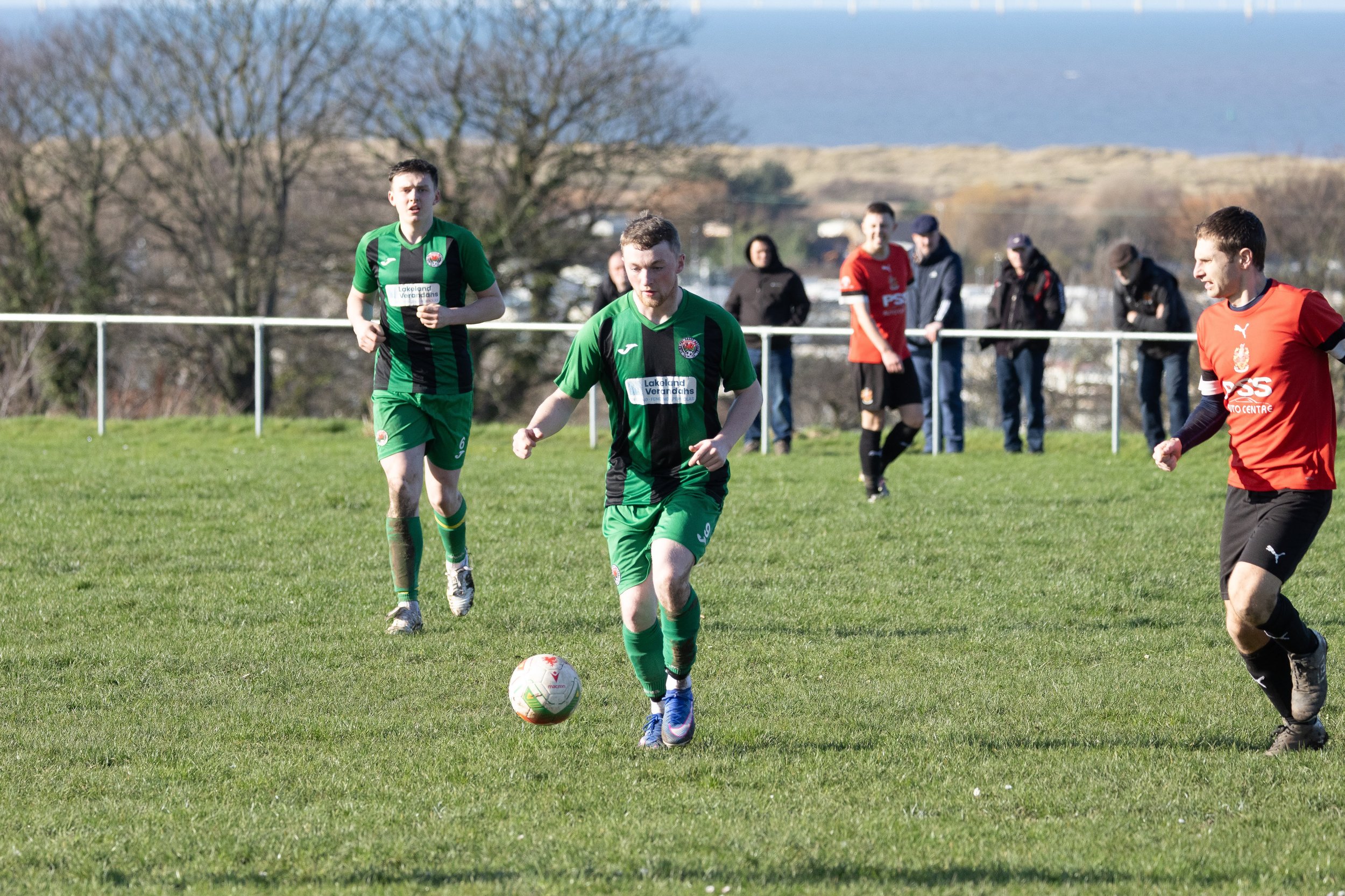 Soccer players in green and red uniforms playing on a grass field during daytime with spectators watching from behind a white fence, trees, and hills in the background.
