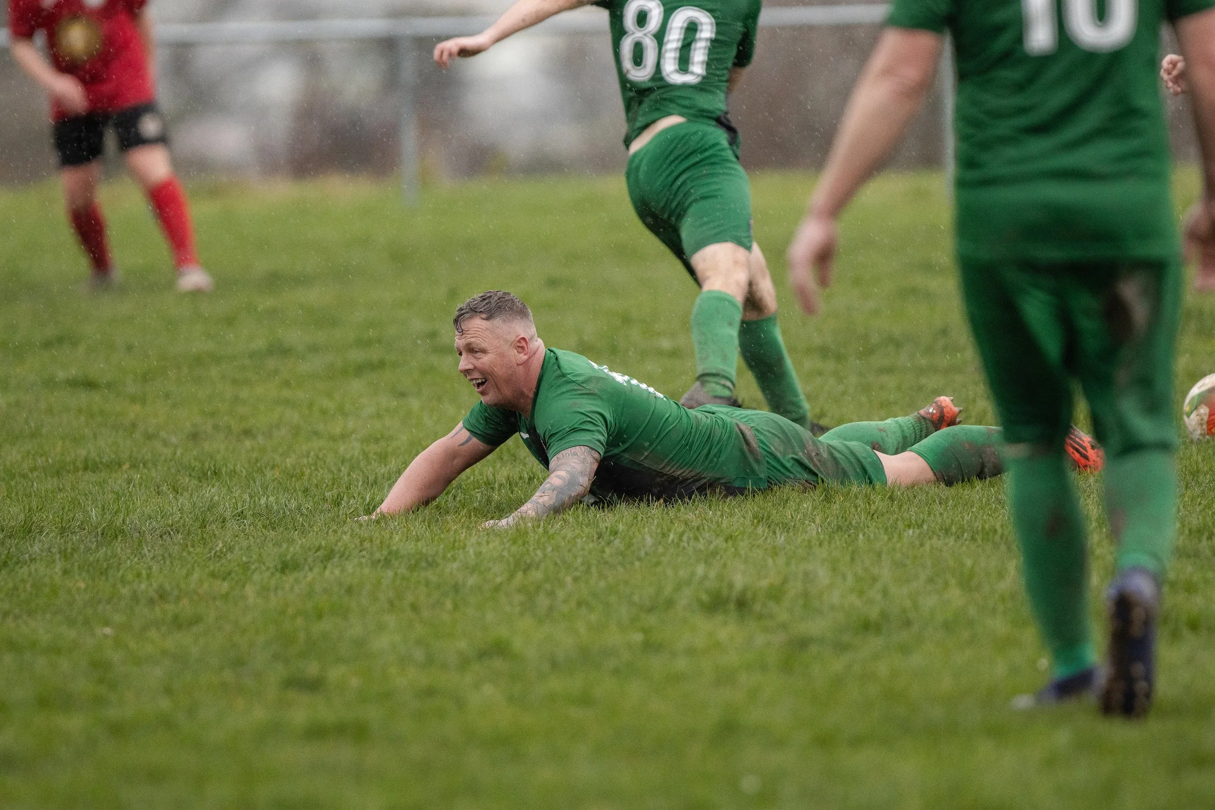 A soccer player in a green uniform lies on the wet grass, smiling after a play, while other players in green and red uniforms are visible around him during a rainy match.