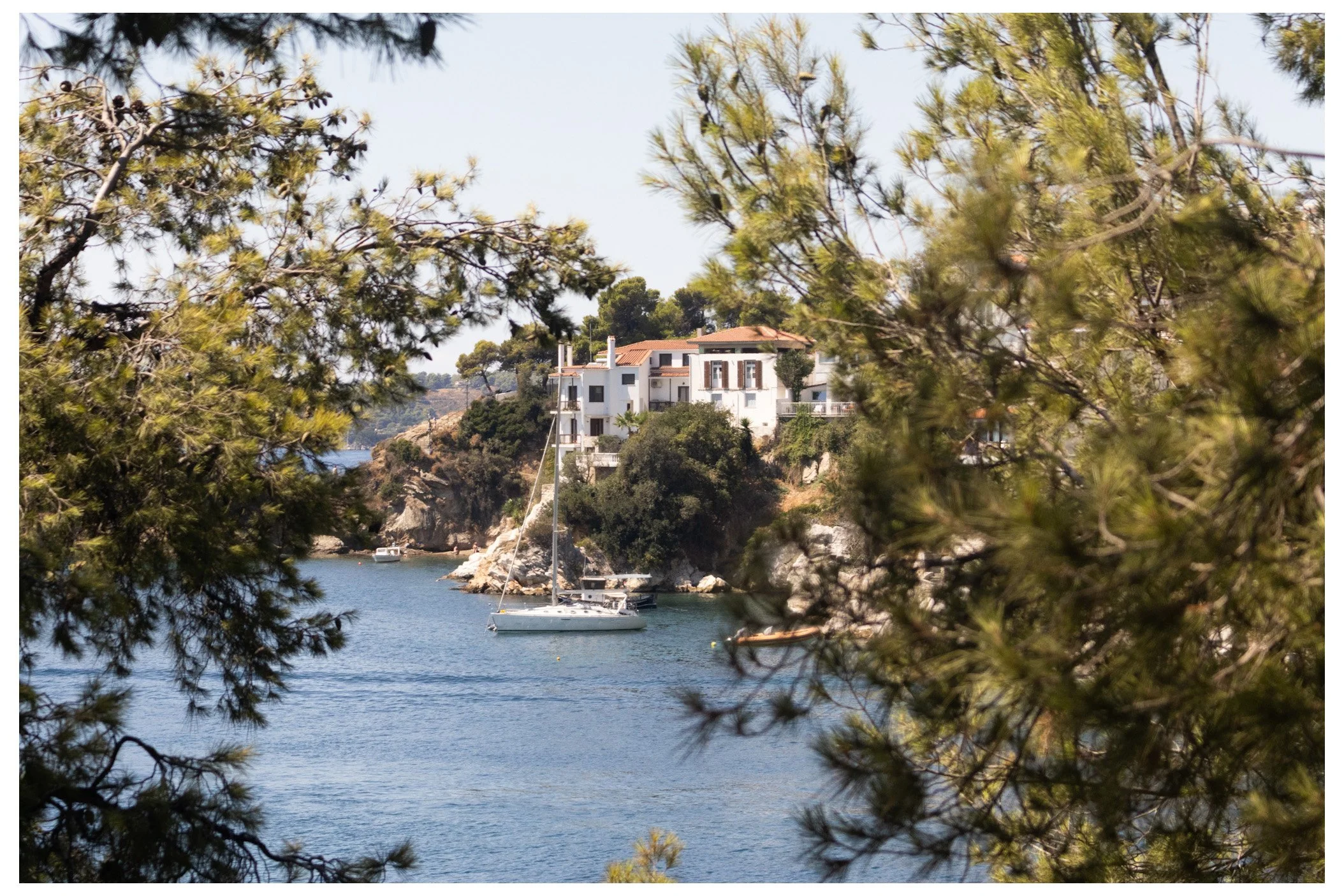 A view of a coastal residential area with white houses on a cliff, seen through trees near a body of water with a sailboat anchored nearby.