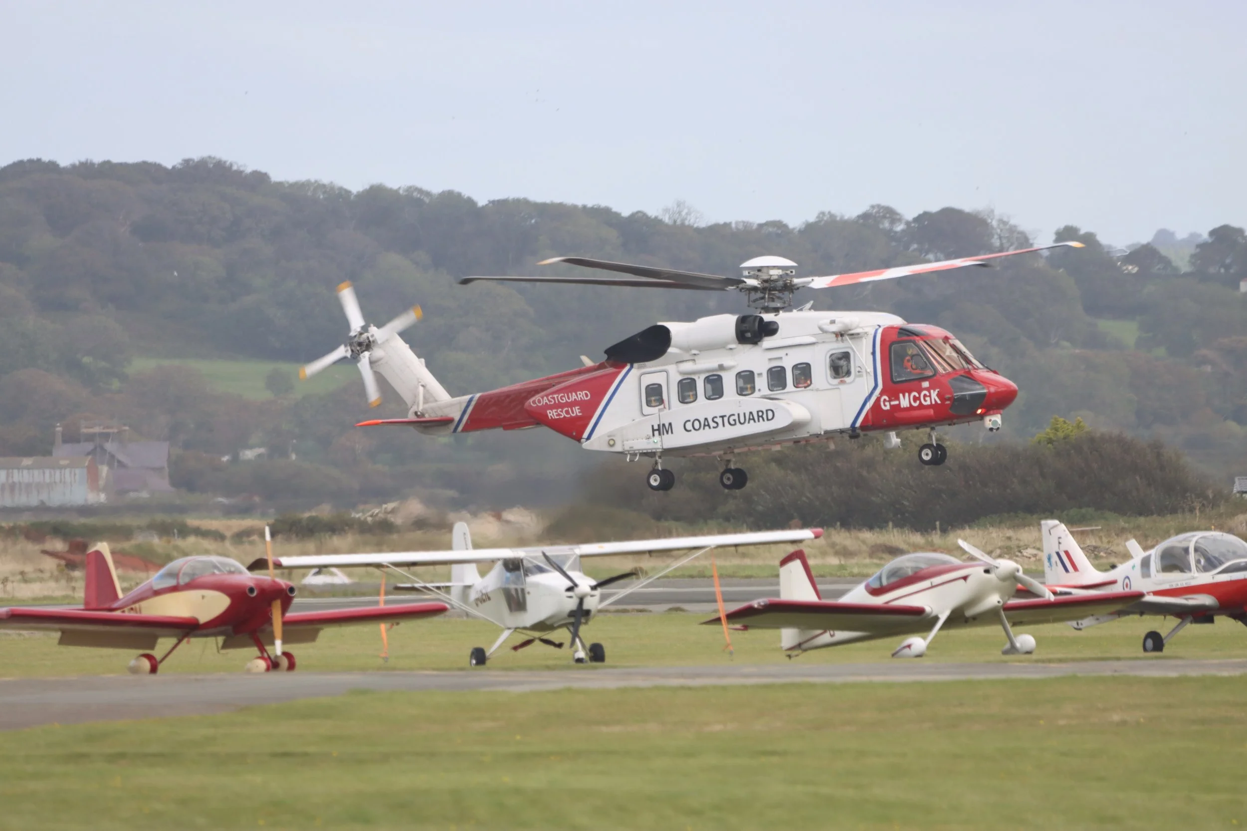 A red and white HM Coastguard rescue helicopter flying low over a field with small planes parked nearby, a grassy hill and trees in the background.