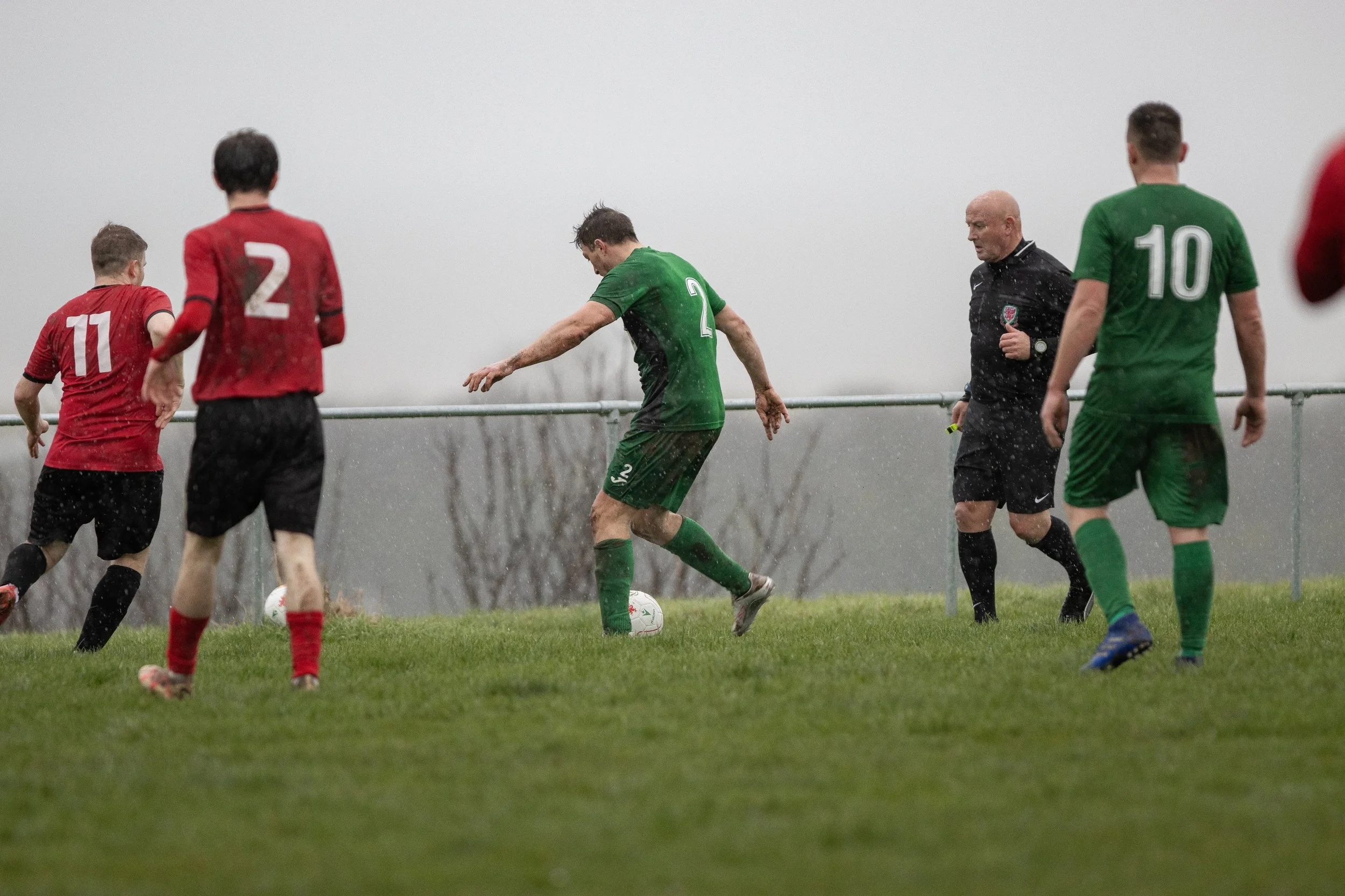 Soccer players and referee on a rainy day during a match, with players in red and green jerseys on a grassy field.