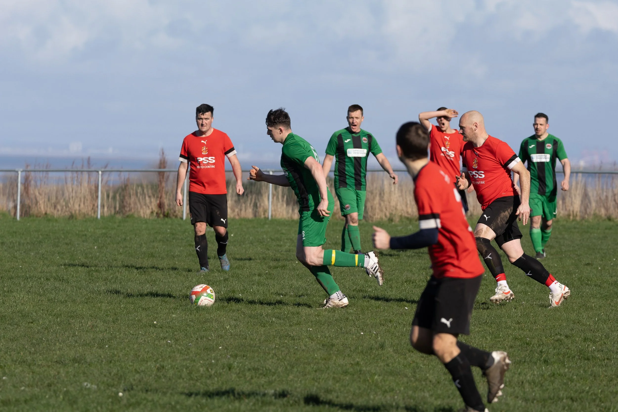 Soccer match with players in red and green uniforms on a grassy field, with a cloudy sky in the background.
