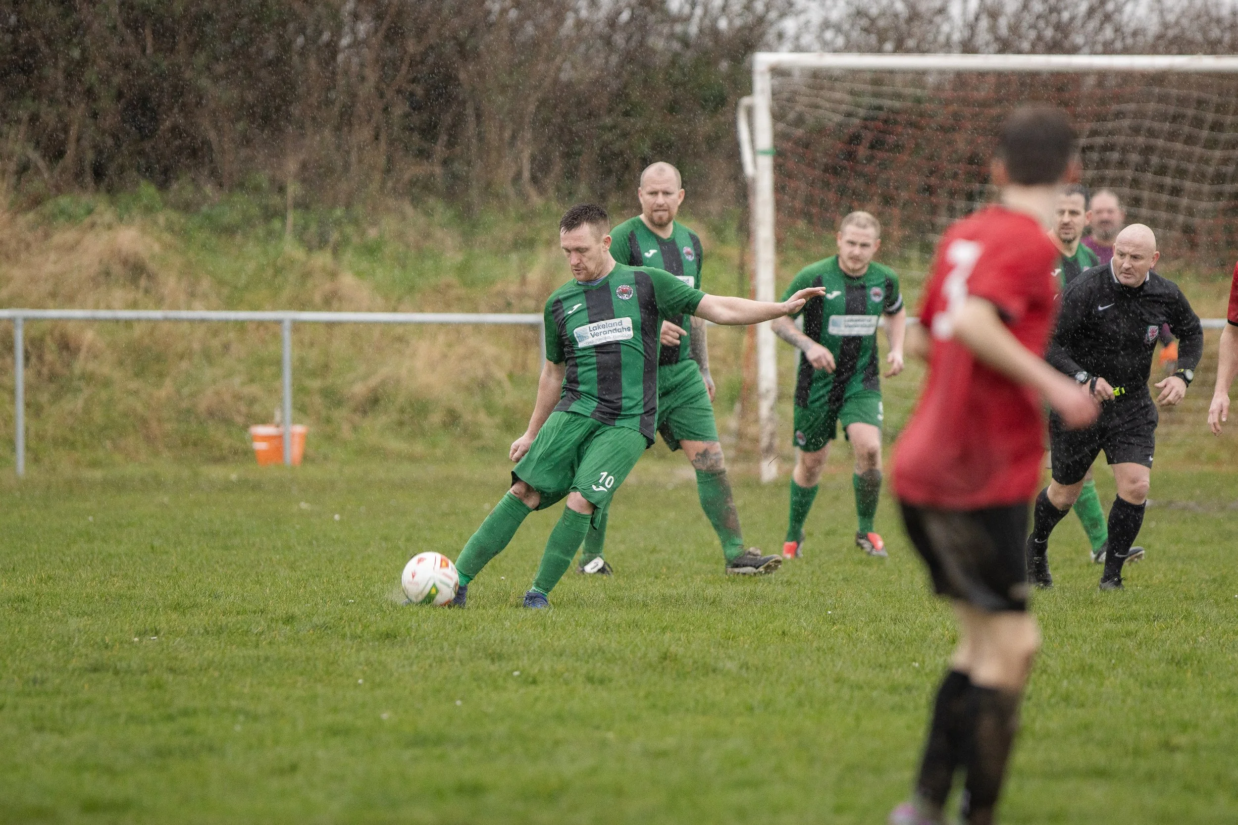 A soccer player in green uniform kicking a soccer ball on the field during a rainy game, with other players and a referee watching in the background.