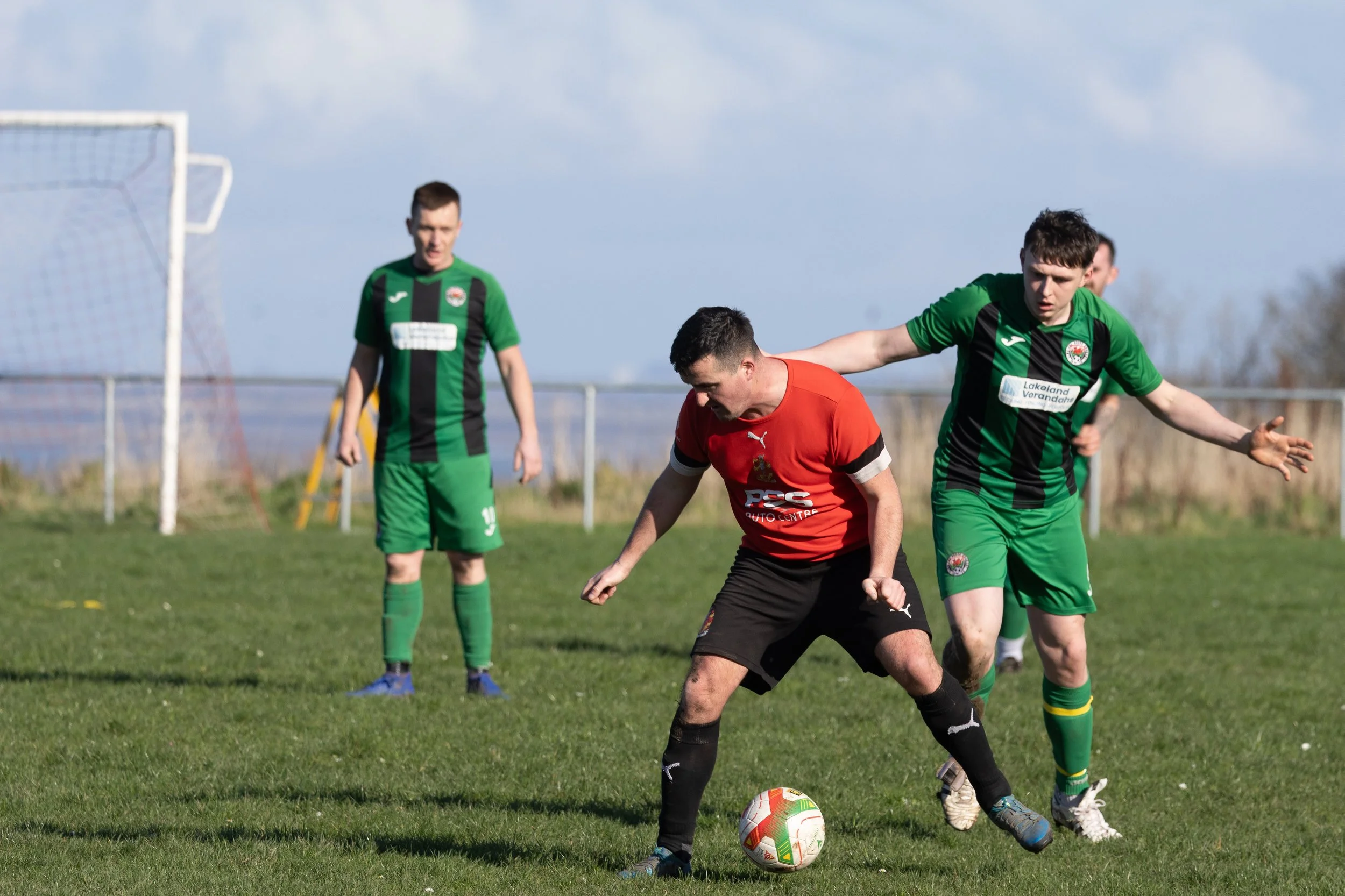 Three soccer players on a grassy field during a match, with one player in a red jersey controlling the ball while two players in green jerseys defend.