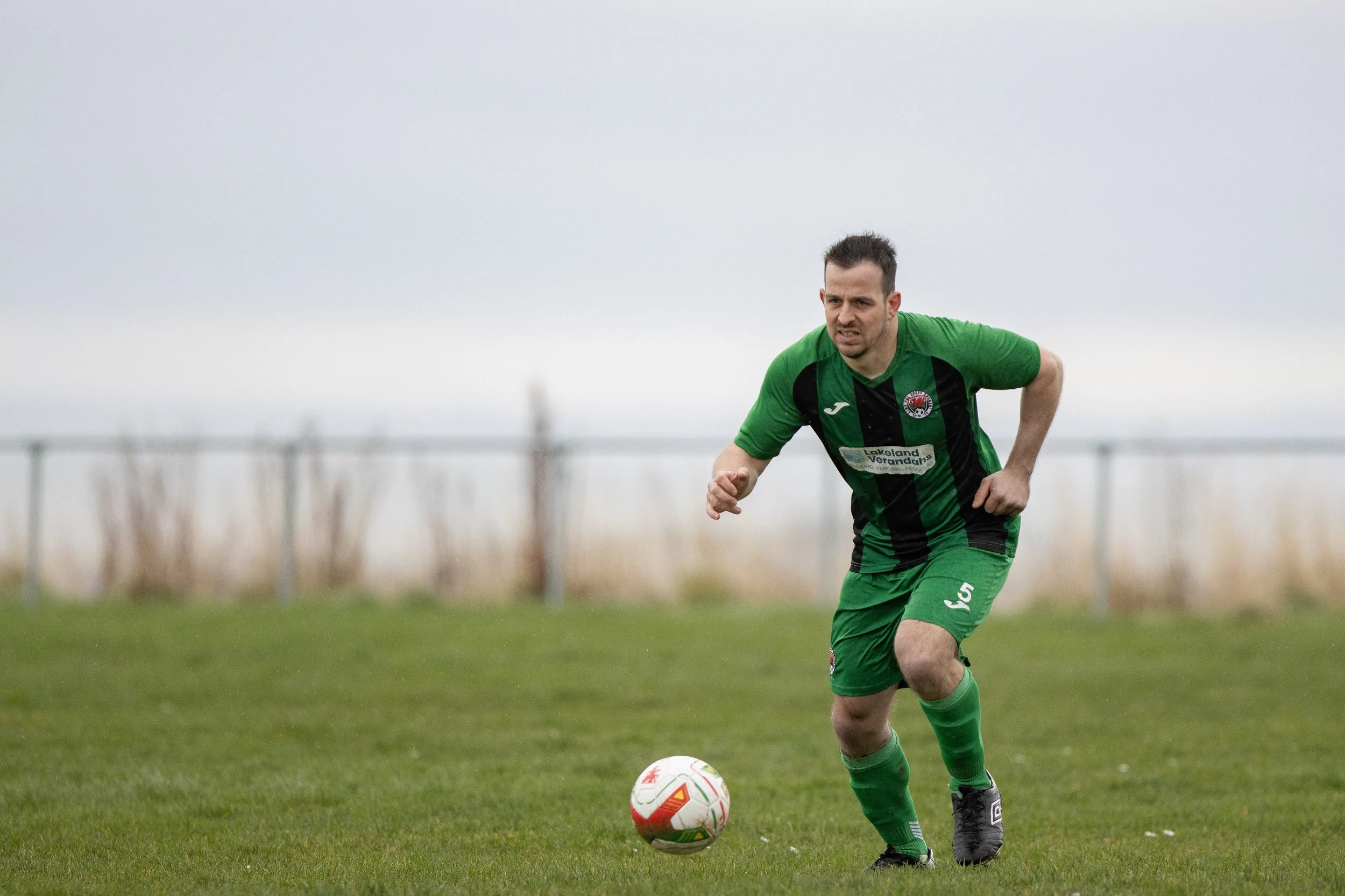 A man in green and black soccer uniform running on a grassy field with a soccer ball.