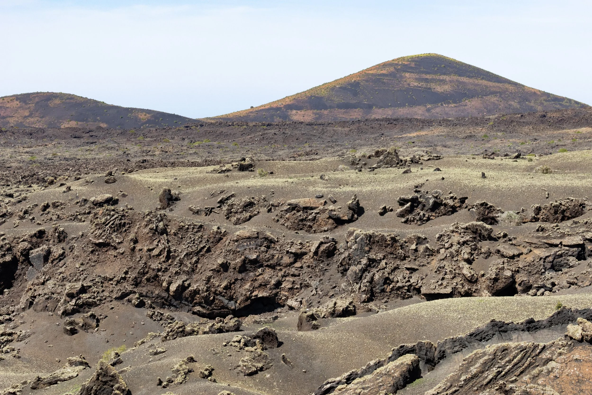A volcanic landscape with dark, rocky terrain and two rounded volcanoes in the background under a clear sky.