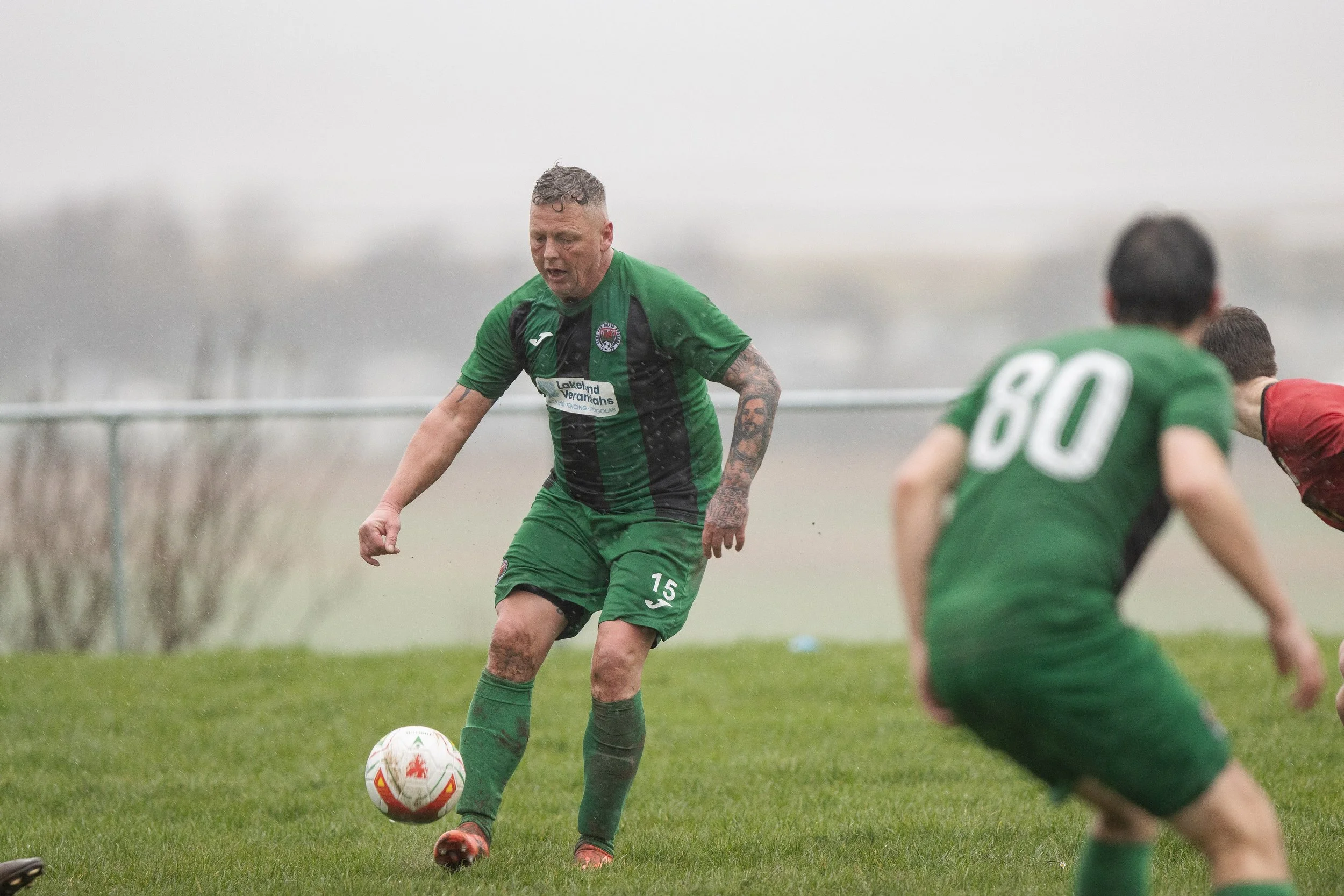 Soccer players competing outdoors in rainy weather, with one player in green controlling the ball.