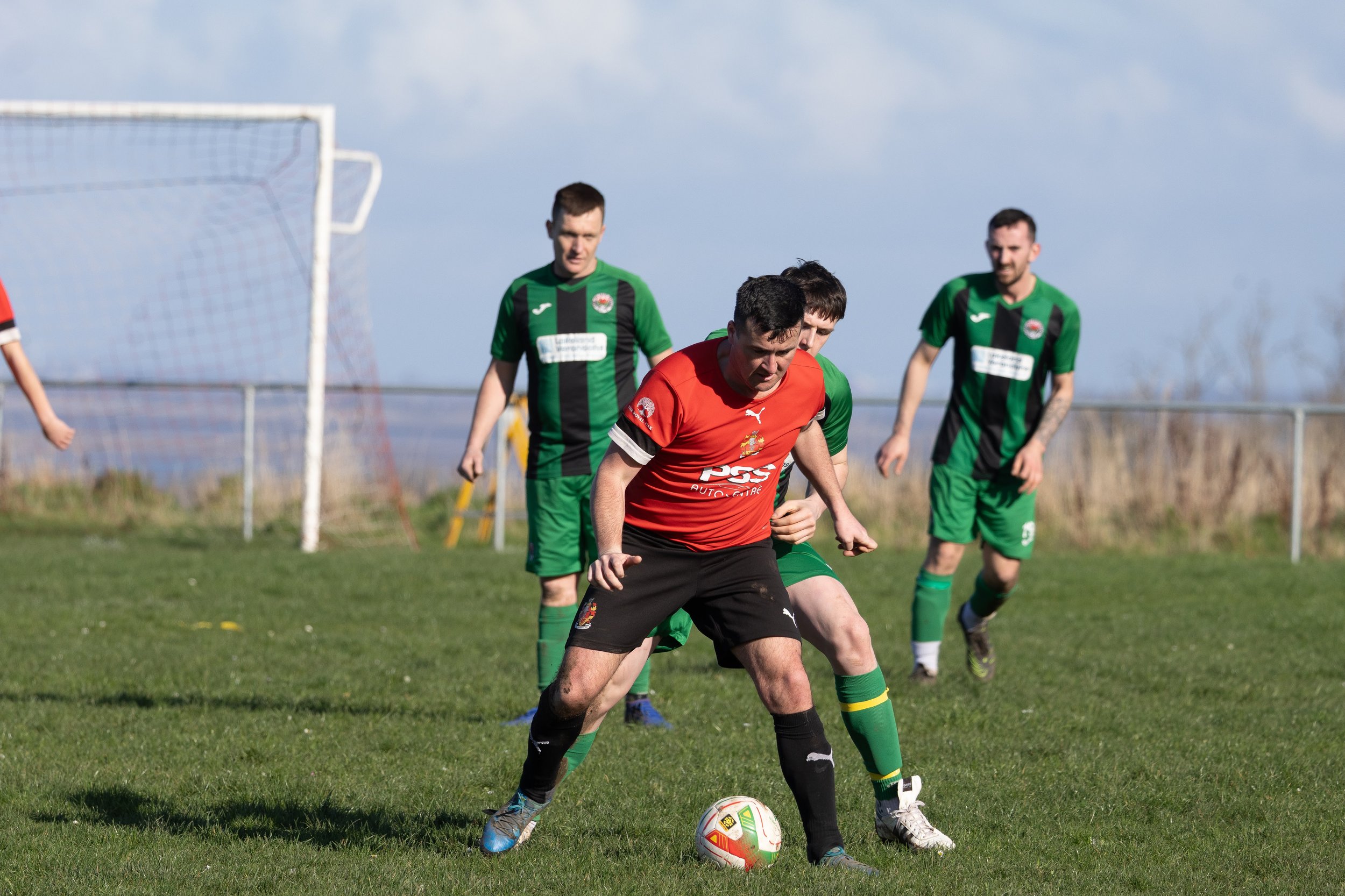 A soccer match with players in green and red jerseys on a grassy field, with a goal post in the background.