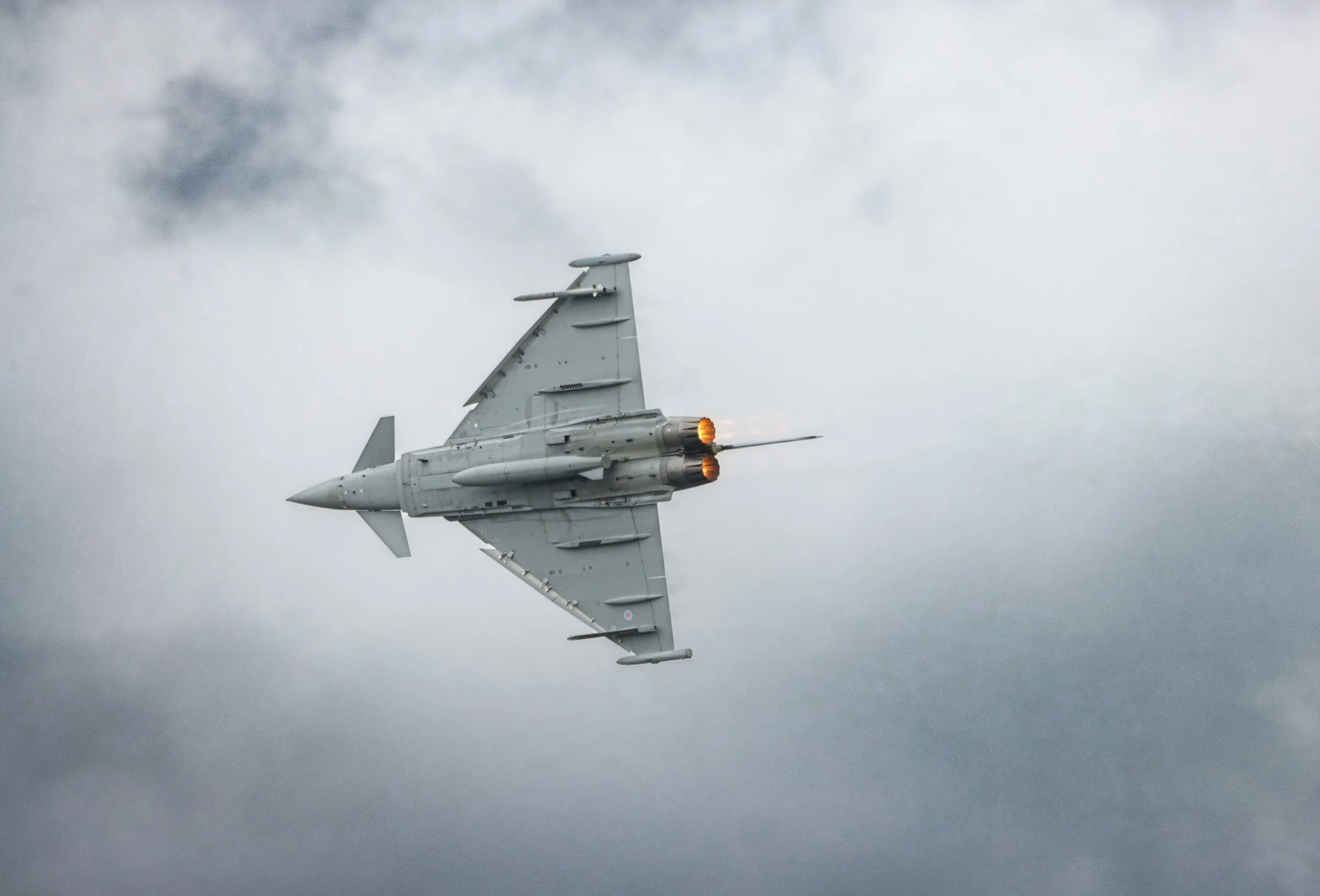 A fighter jet airplane flying through the cloudy sky with its afterburners on.