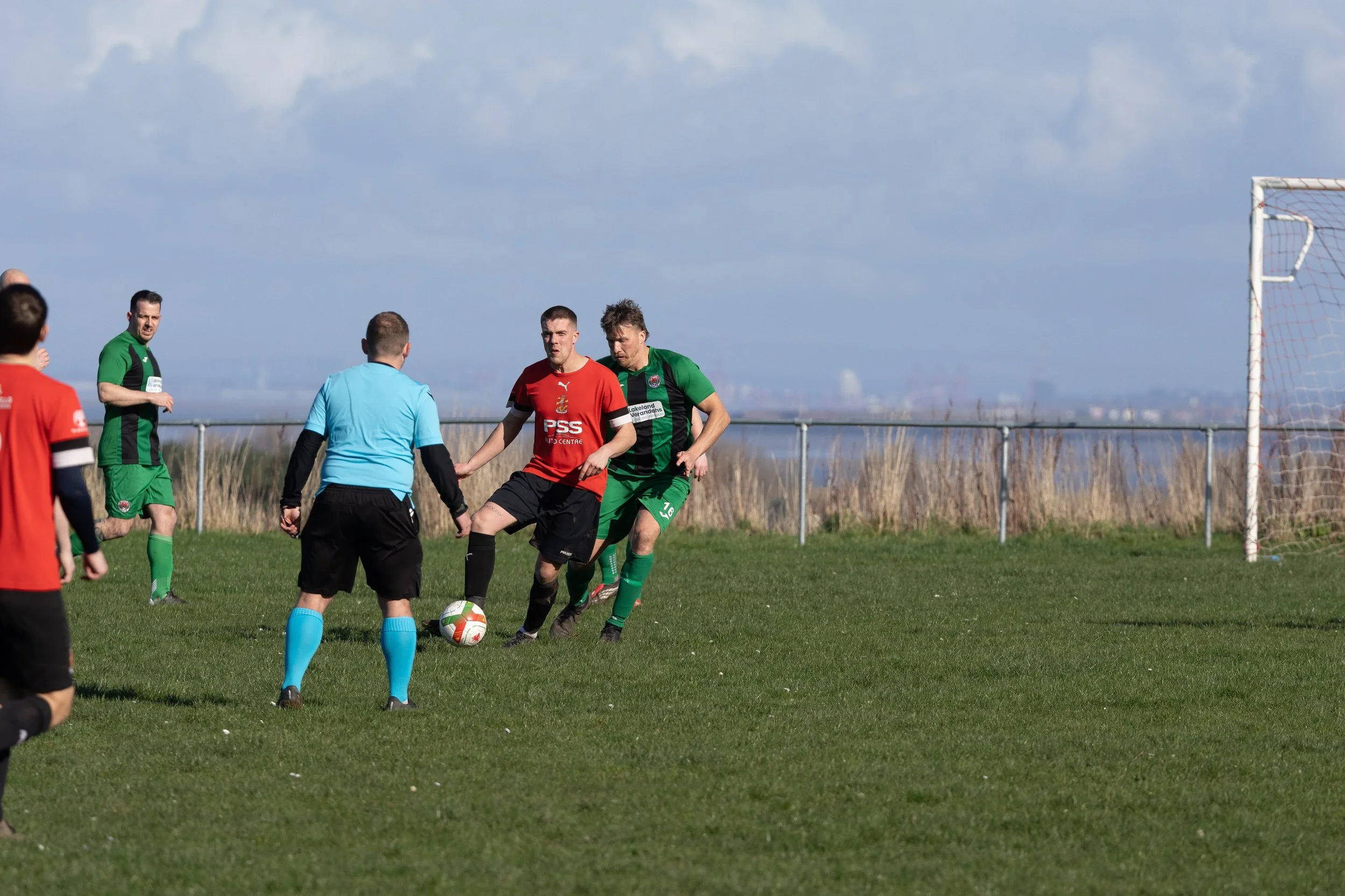 Soccer players and referee on the field during a match with a goalpost and fence in the background.